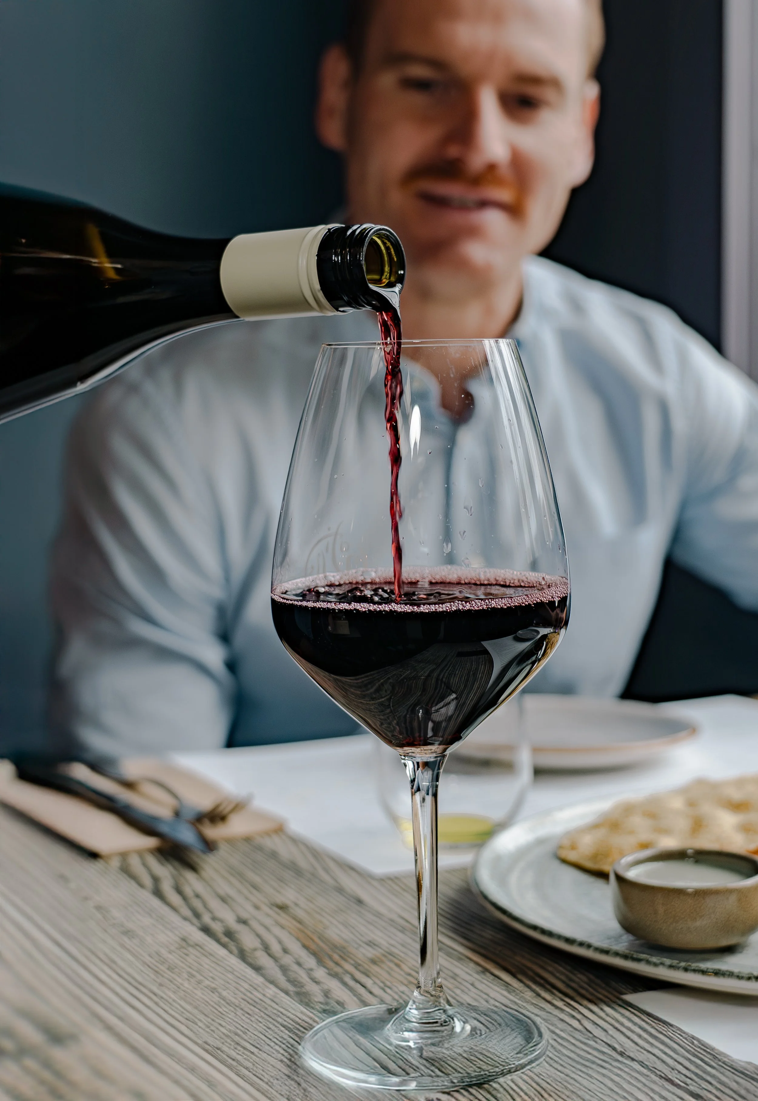 A man with a moustache & red wine being poured into a tall wine glass at a dining table.
