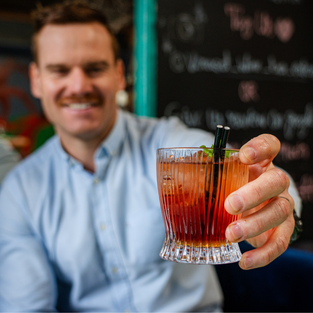 A man in a light blue shirt holding a glass of red cocktail with black straws in a bar or restaurant setting.