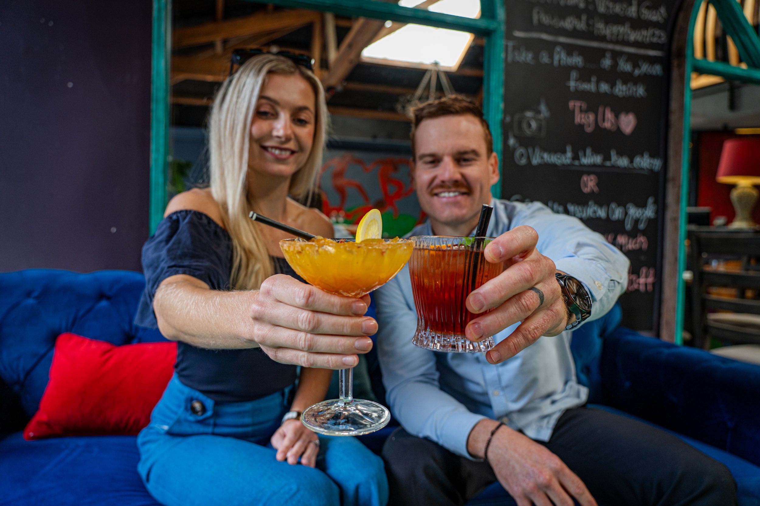 A woman with blonde hair and a man with brown hair sitting on a blue couch, smiling, holding drinks in glassware with lemon slices, in a cozy restaurant with a chalkboard menu in the background.