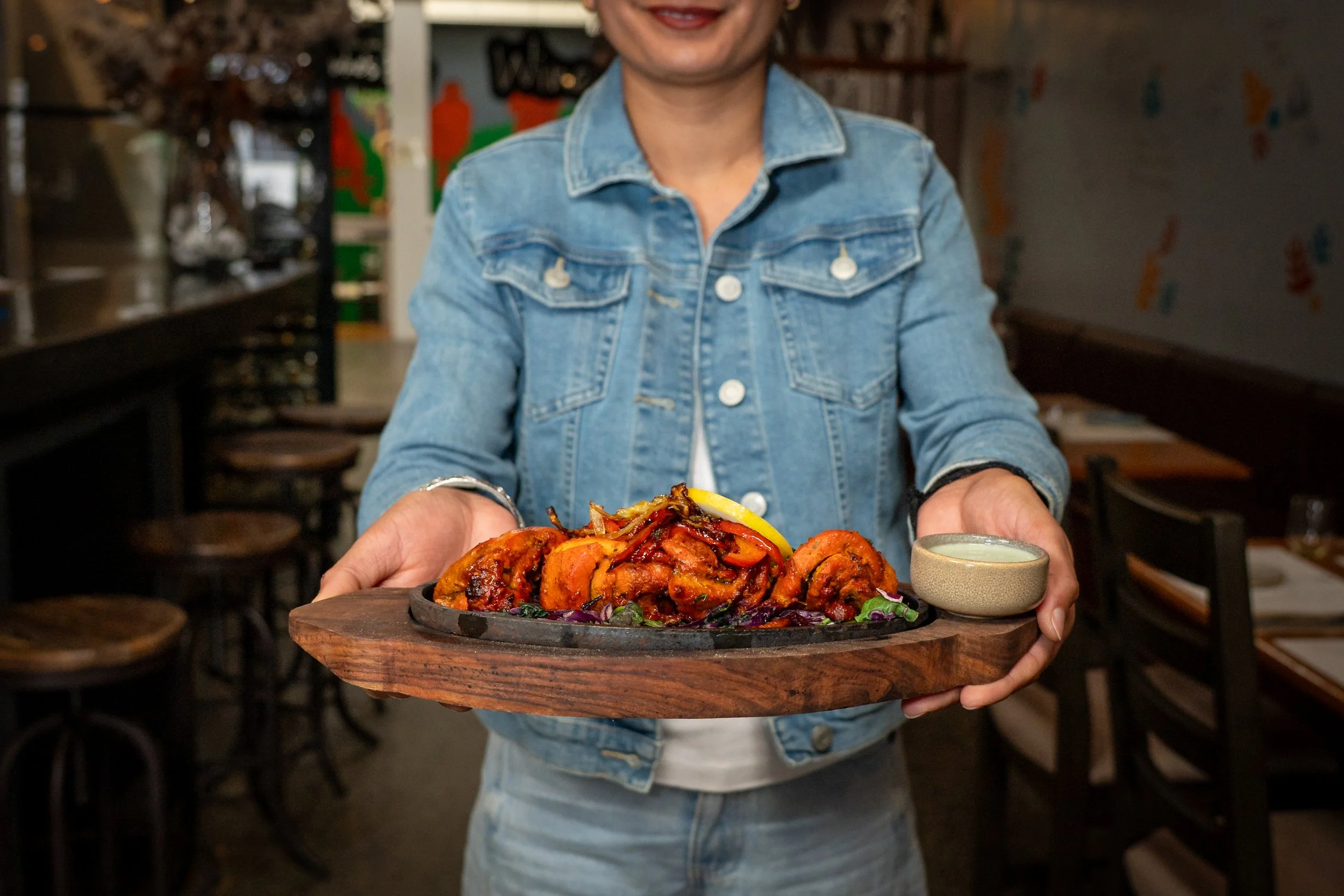 A person in a denim jacket is holding a wooden tray with a large serving of spicy Korean-style grilled chicken and vegetables, along with a small bowl of dipping sauce in a restaurant setting.