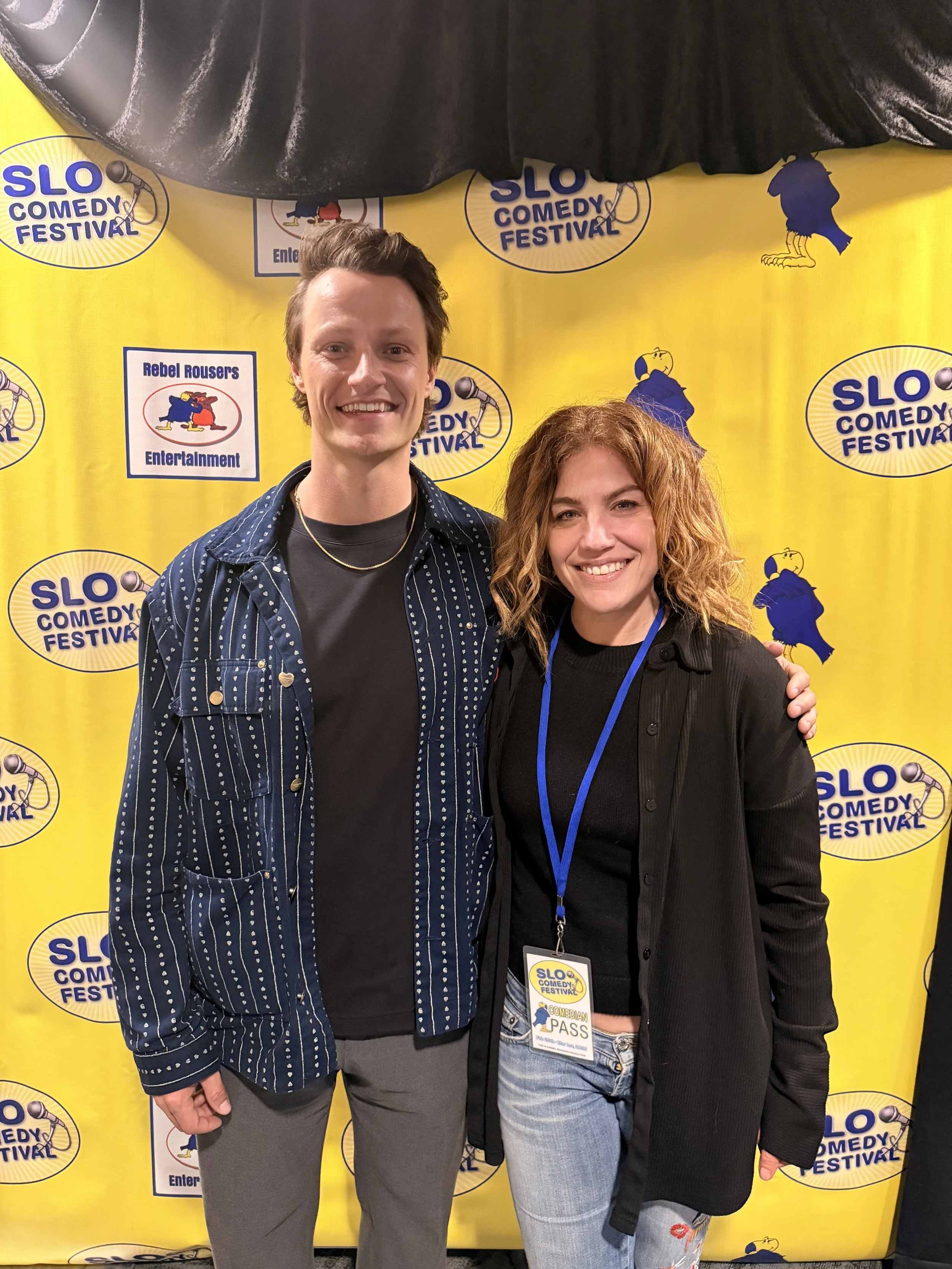 Two people standing in front of a yellow backdrop with the 'SLO Comedy Festival' logo, smiling at the camera. One is wearing a dark striped shirt with a gold necklace, and the other is wearing a black jacket with a lanyard and pass badge.