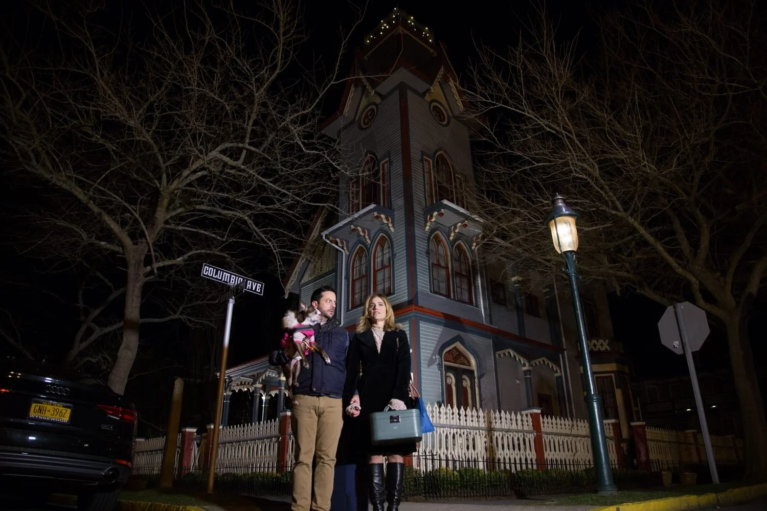 A man and woman stand on a sidewalk at night in front of a Victorian-style house, with the man holding a small dog and the woman carrying a suitcase and a shopping bag. There is a street sign that reads 'Columbic Ave' and a street lamp.