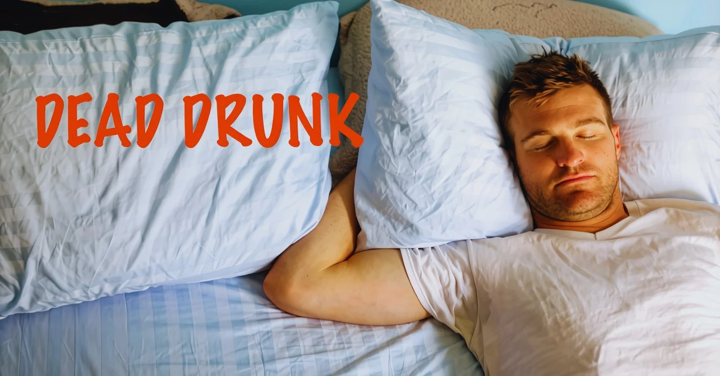 A young man sleeping on a bed with white sheets, resting his head on a pillow, with a pillow with the words 'DEAD DRUNK' written in red letters nearby.