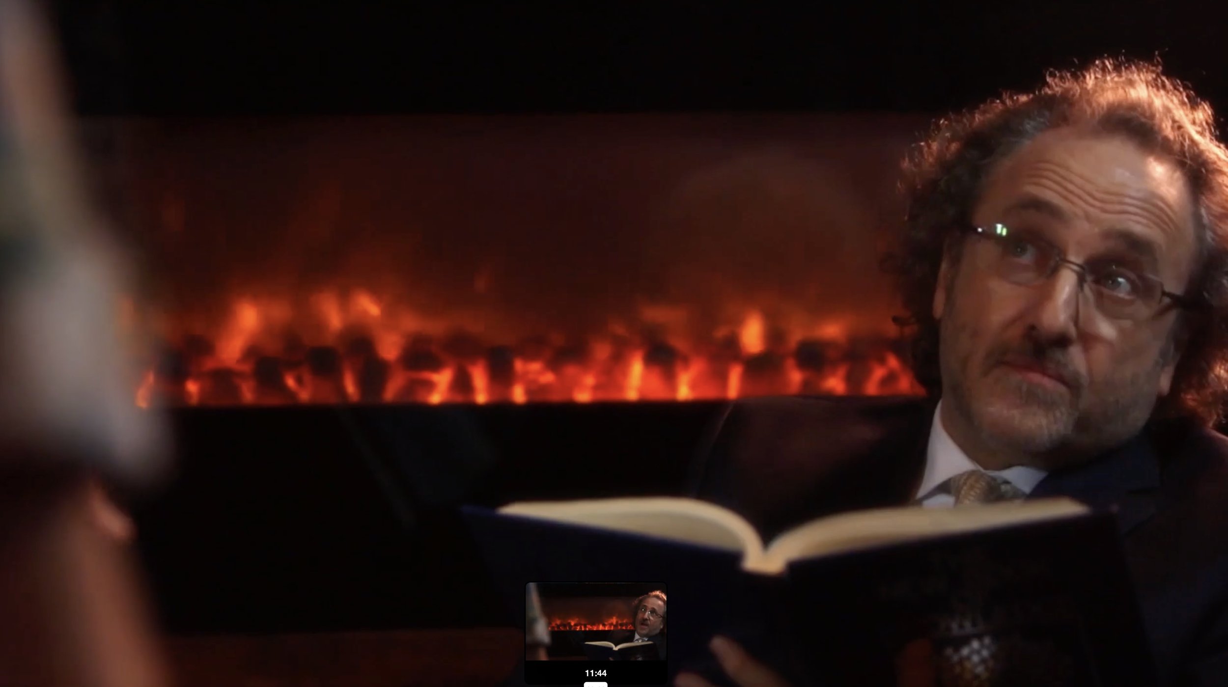 A man with glasses and curly hair sitting in a car at night, with a fire seen through the rear window.