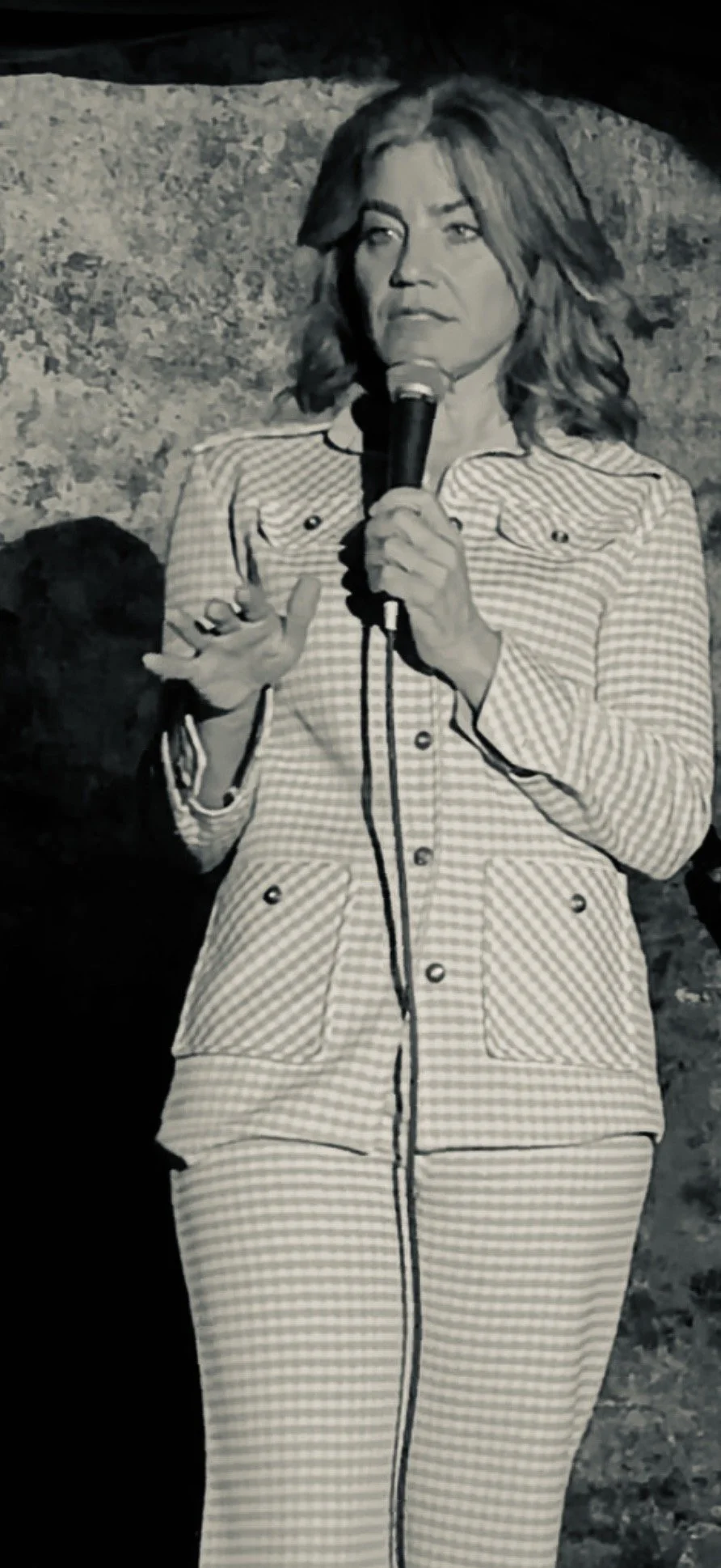 Black and white photo of a woman holding a microphone and speaking, wearing a checkered outfit, standing against a rocky background.