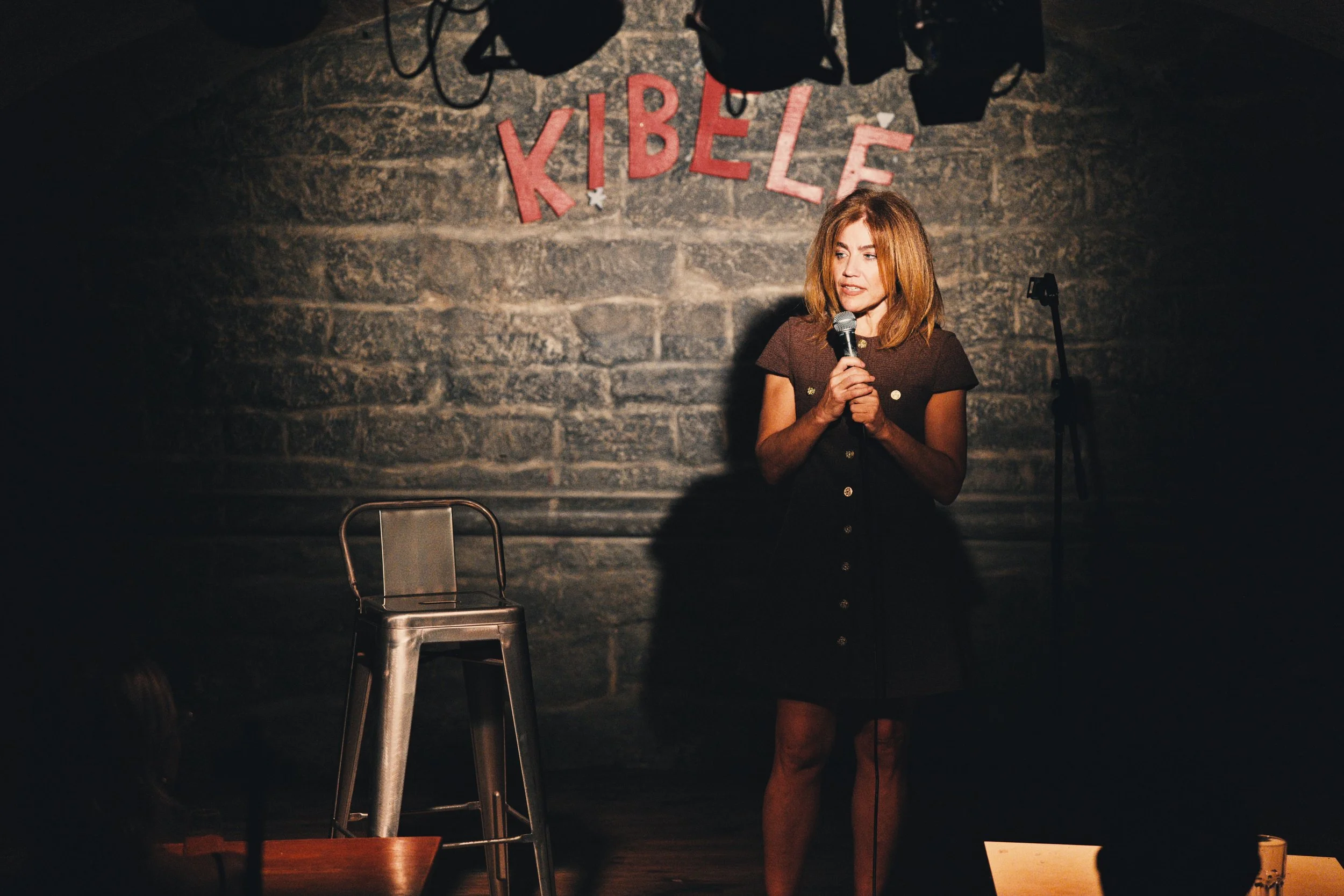 Dana Marisa holds a microphone on stage in front of a brick wall with red letters spelling 'KRBEL'. She appears to be speaking or performing, and there is a metal chair in front of her. The stage is dimly lit.