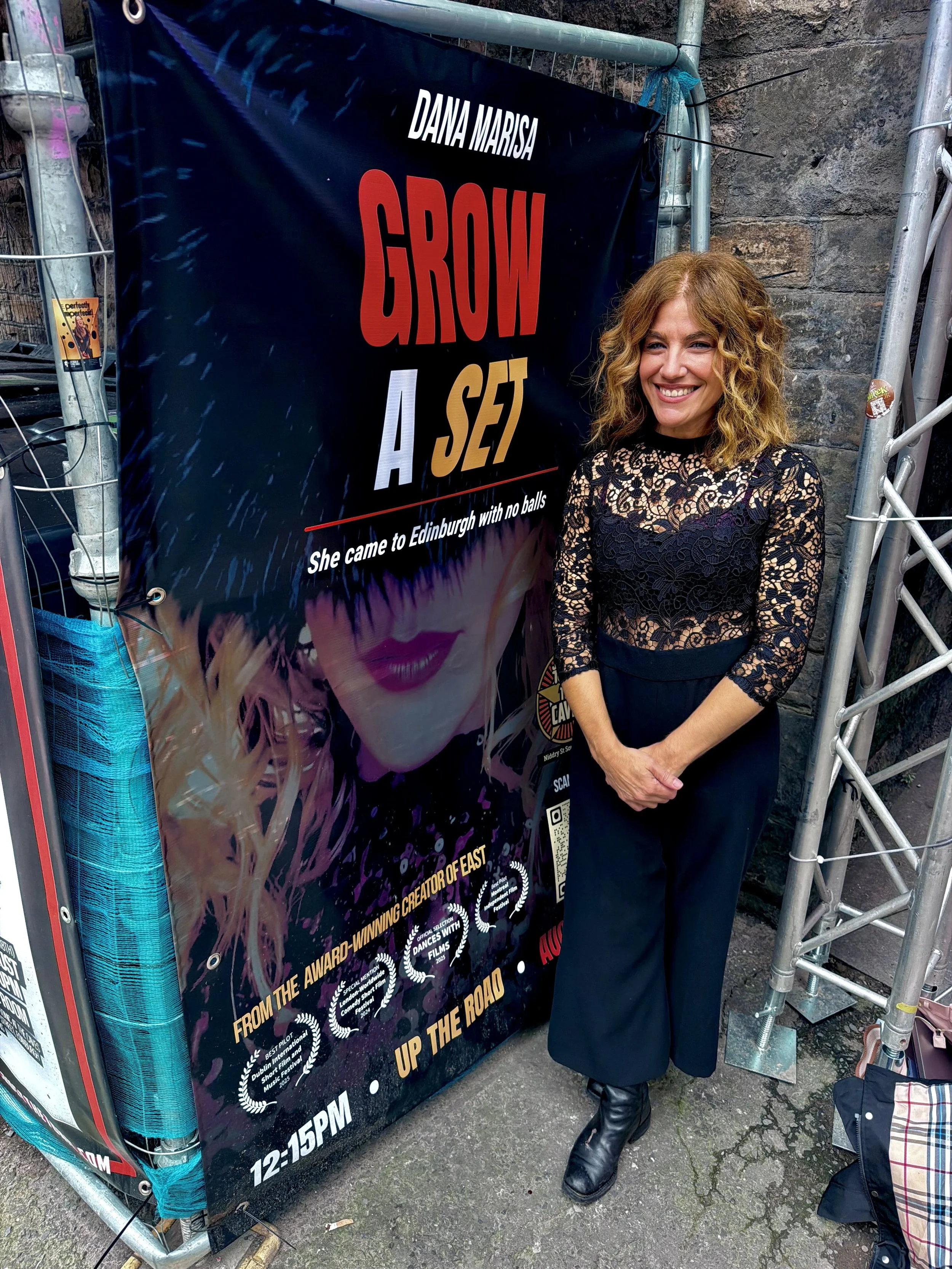 A woman with curly red hair wearing a black lace top and dark pants, standing next to a large poster with bold text, in an outdoor setting with a stone wall and metal scaffolding.