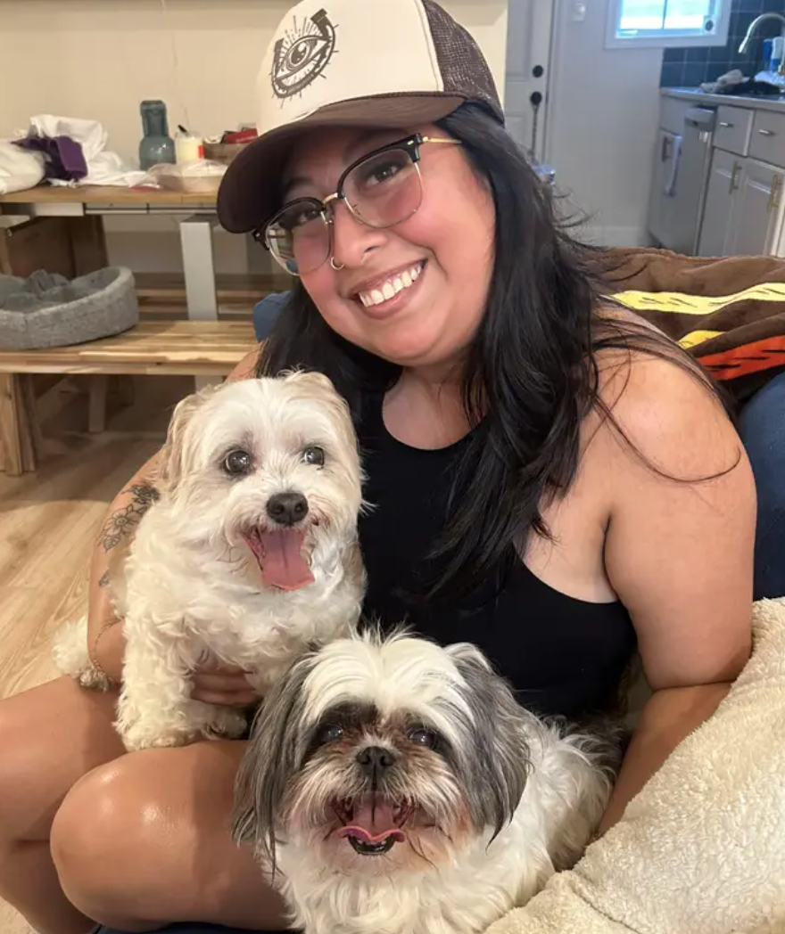 A woman with glasses and a cap smiling while hugging two small dogs, one with white curly fur and the other with black and white fur, inside a cozy home.