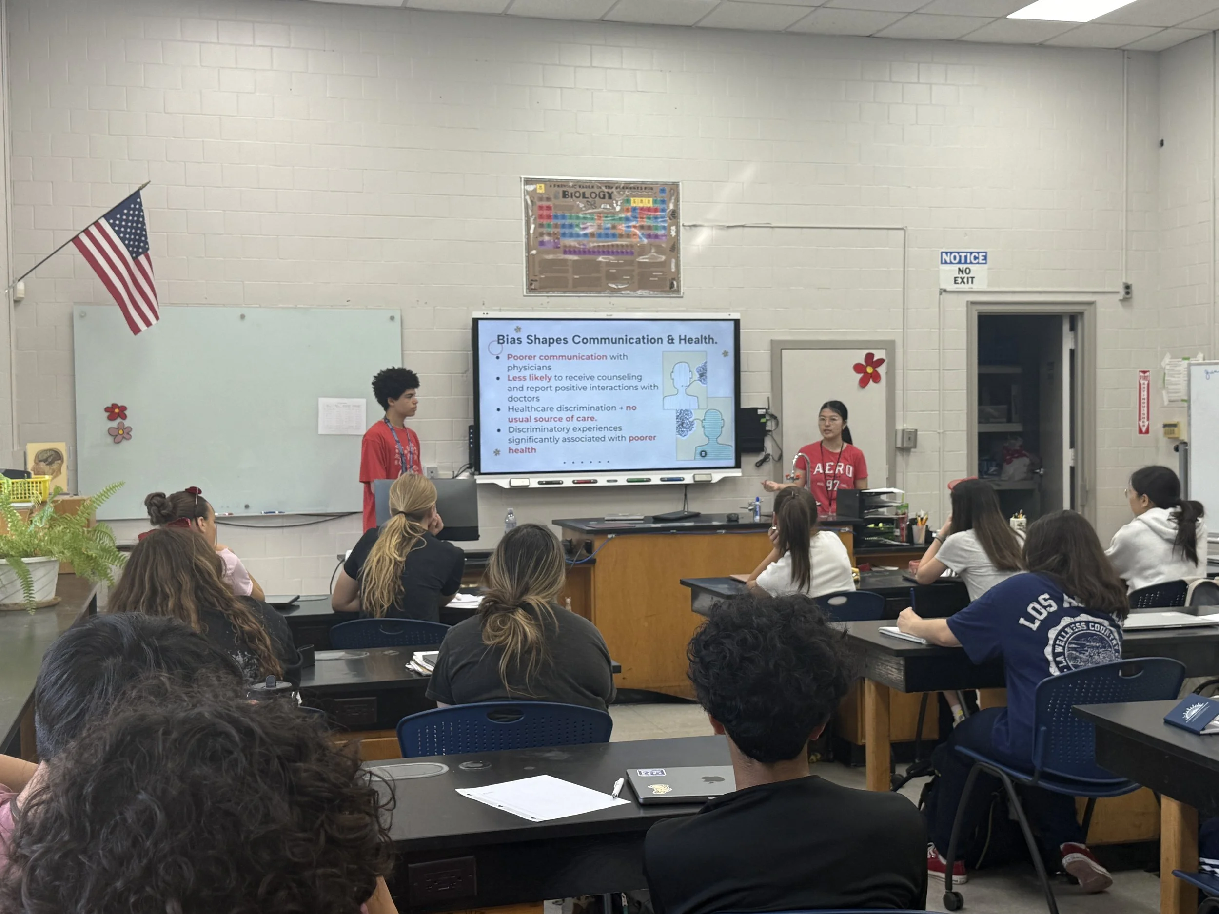 A classroom with students seated at desks facing a screen that displays a presentation slide titled 'Bias Shapes Communication & Health.' Two students are standing at the front, one speaking and the other listening. The classroom has a white dry-erase board, an American flag, and various decorations on the walls.