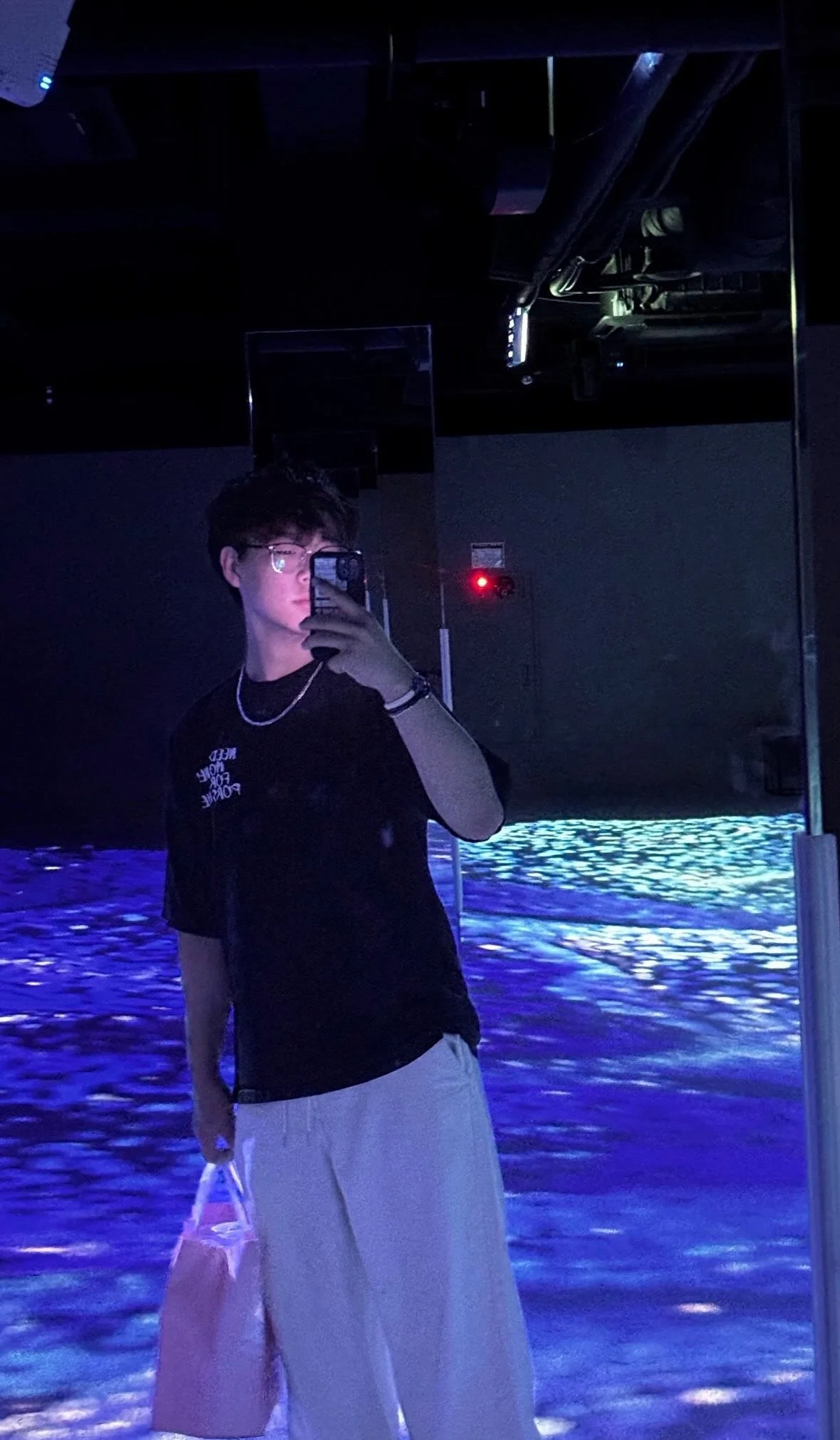 A person taking a mirror selfie with a smartphone in a dark, indoor setting with blue and purple lighting, reflective elements, and water-like patterns on the floor.