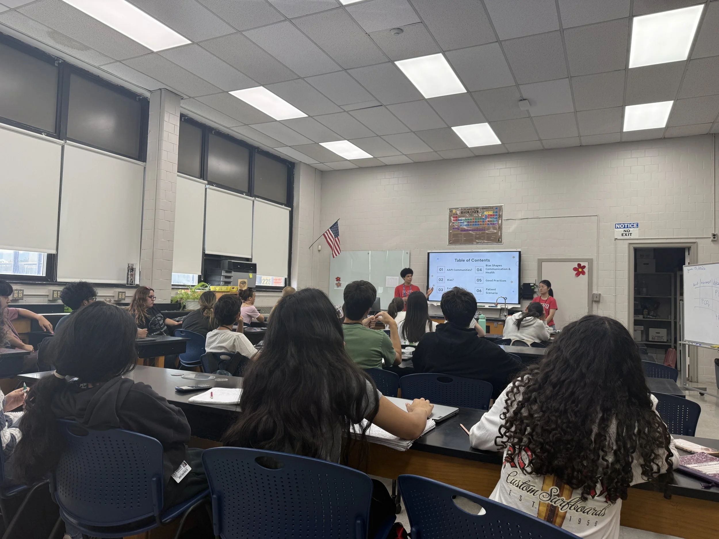 A classroom filled with students seated at desks facing a large screen at the front of the room. A female teacher is standing near the screen, giving a presentation. The classroom has high ceilings with bright fluorescent lighting, and an American flag is displayed near the windows. The room has white brick walls with posters and notices.