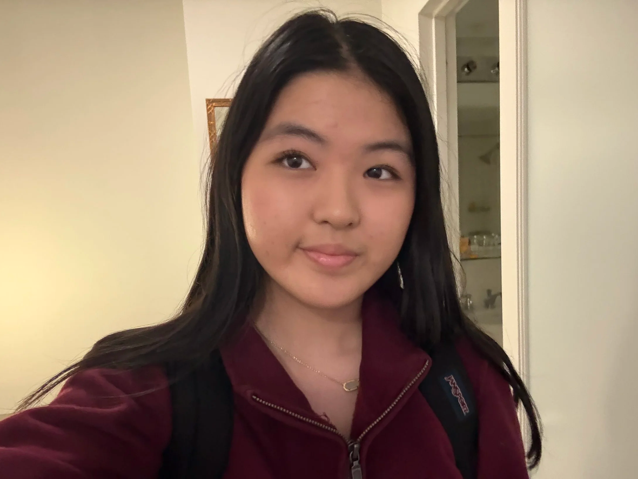A young woman with long dark hair, wearing a maroon jacket and a black backpack, standing indoors with a neutral expression.