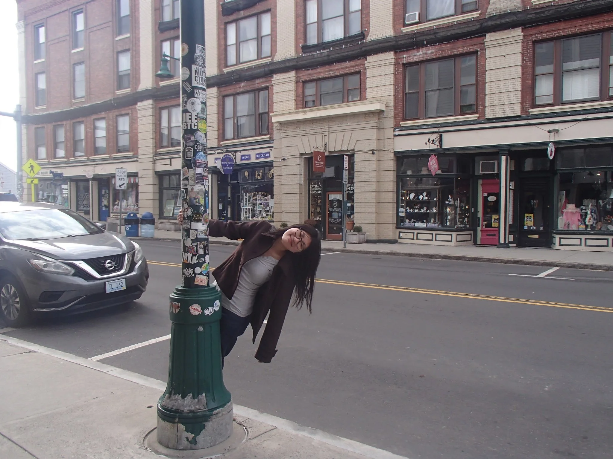 A woman wearing glasses and a dark jacket is leaning over a green trash can covered in stickers, on a city street with cars parked and a row of brick buildings with windows and storefronts in the background.