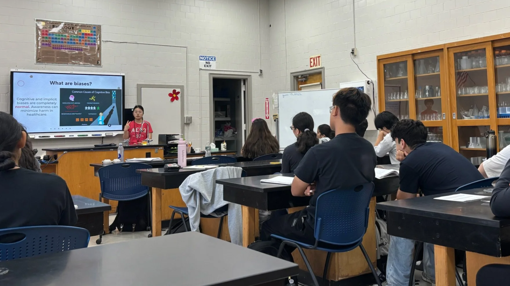 A classroom with students sitting at desks, facing a teacher who is giving a presentation on biases. The presentation slide on the screen discusses common causes of cognitive bias.