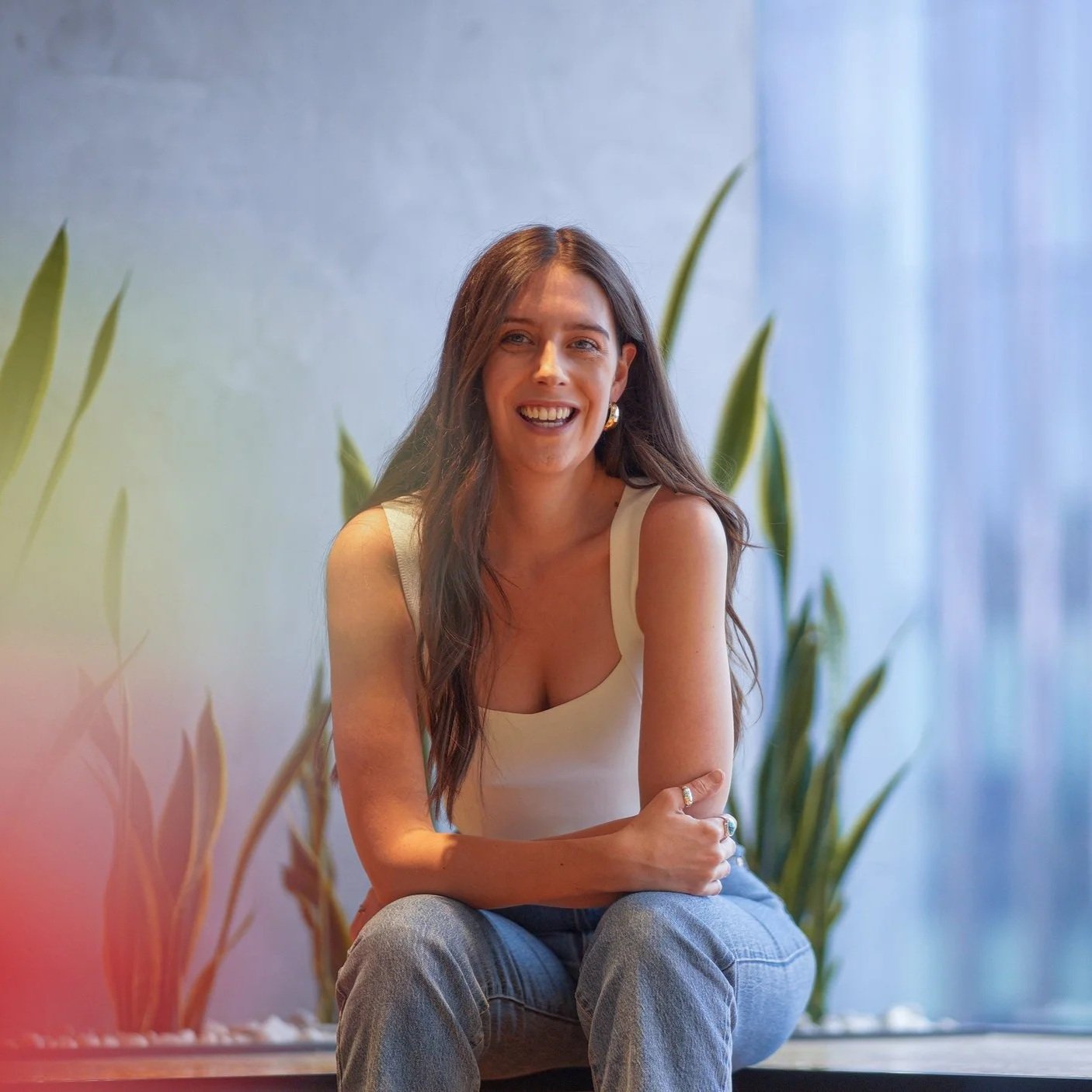 A woman with long brown hair, wearing a sleeveless white top and jeans, sitting with her arms crossed and smiling, with green plants and a blue curtain in the background.