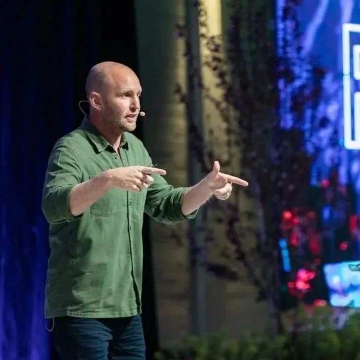 A man in a green shirt giving a presentation on stage, gesturing with his hands.