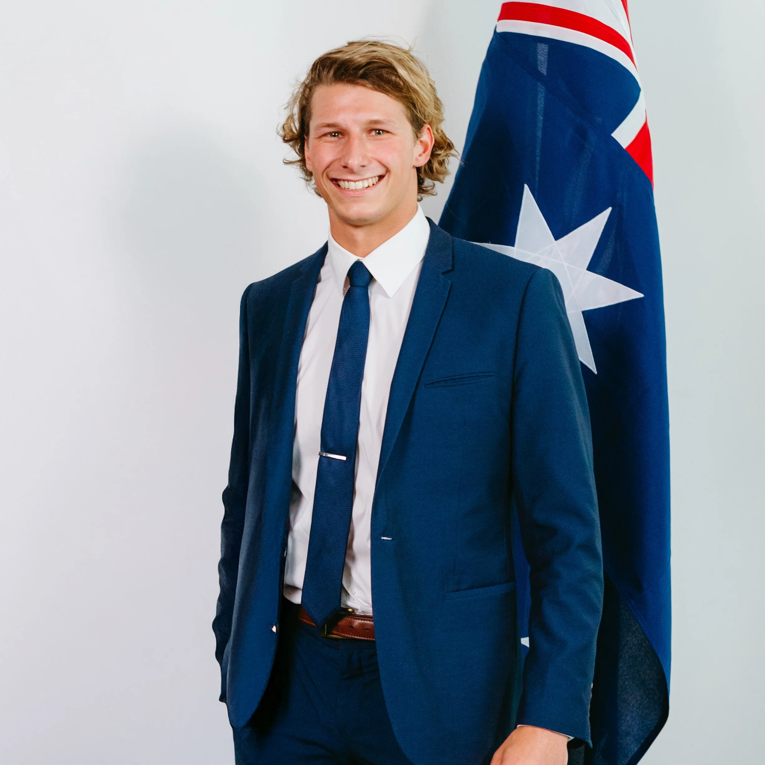 A man with wavy blonde hair smiling, dressed in a blue suit, white shirt, and navy tie, standing in front of a partially visible American flag.