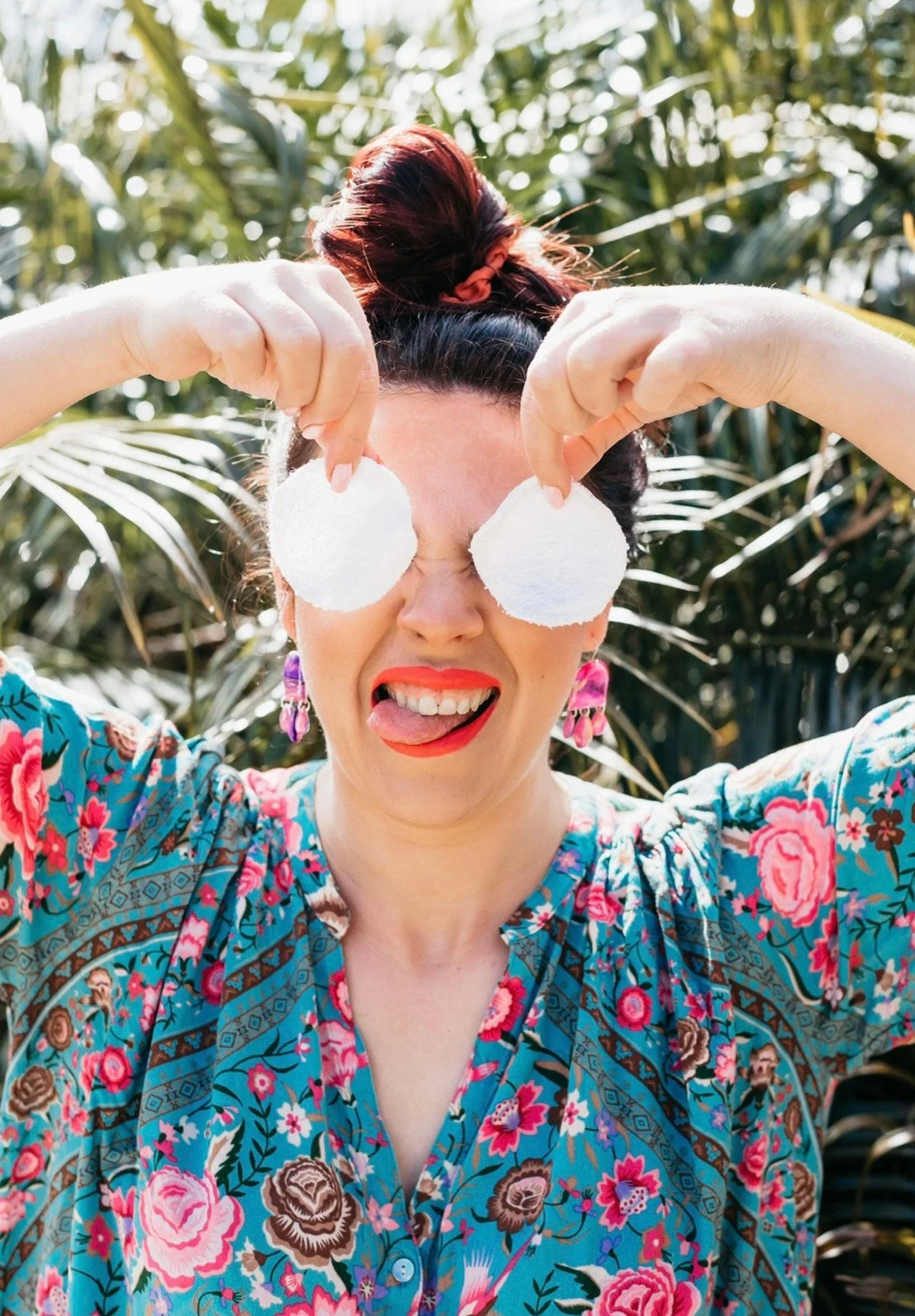 A woman with red hair in a bun holding two large cotton swabs over her eyes, making a silly face with her tongue out, wearing a colorful floral shirt, standing outdoors in front of green plants.