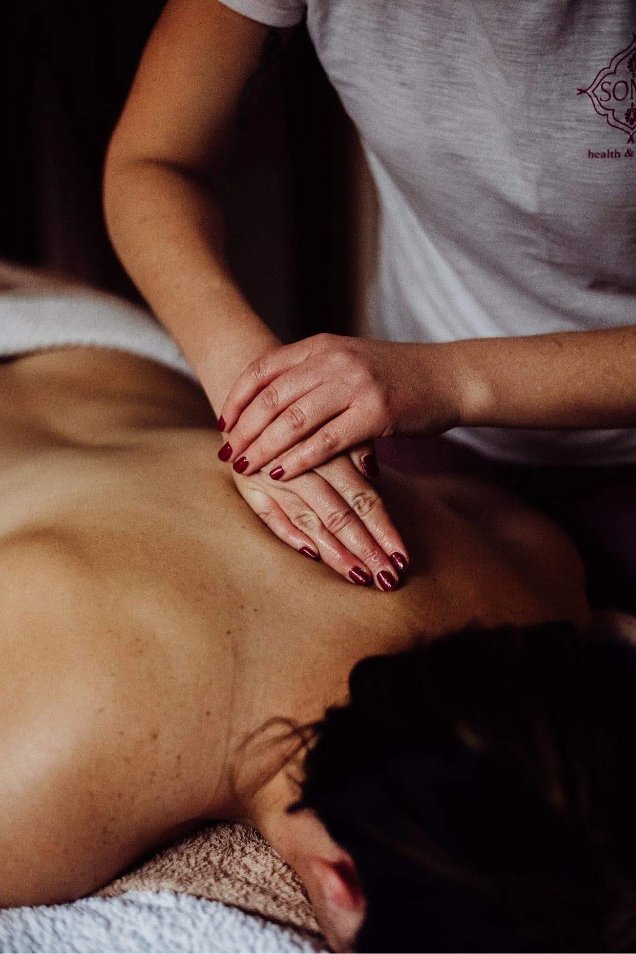 A person receiving a massage on their back from a massage therapist. The therapist has red painted nails and is wearing a gray shirt.