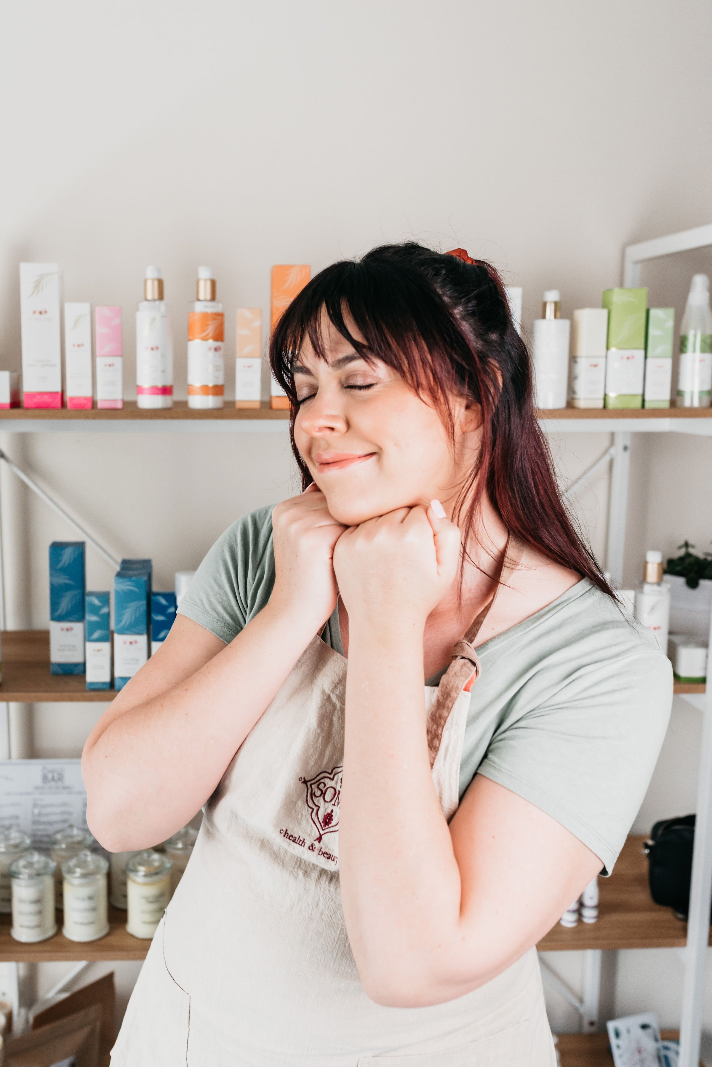 Woman smiling with eyes closed, wearing an apron, in front of shelves with skincare or cosmetic products.