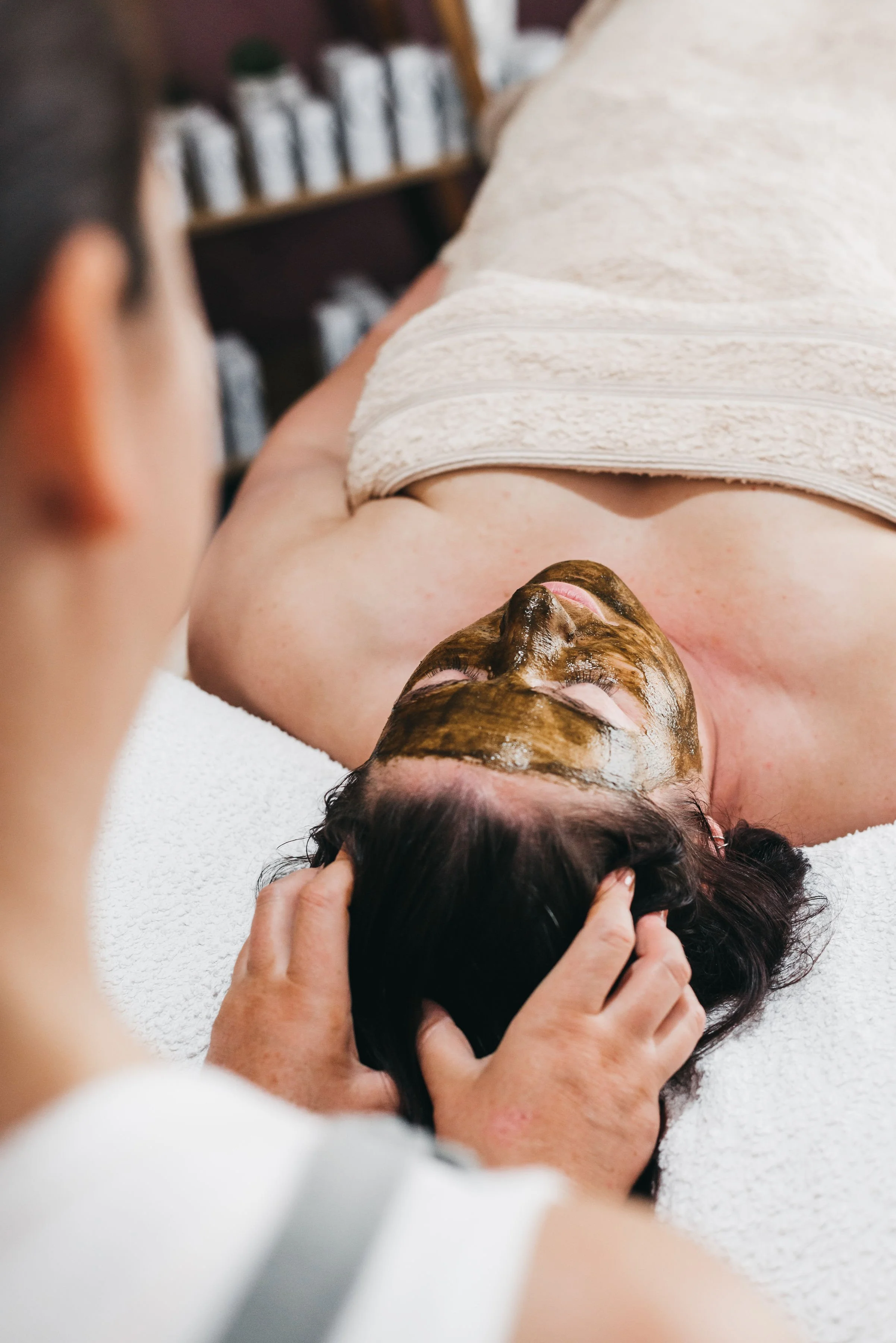 A woman with facial mud mask lying on a towel while receiving a face treatment at a spa or clinic.