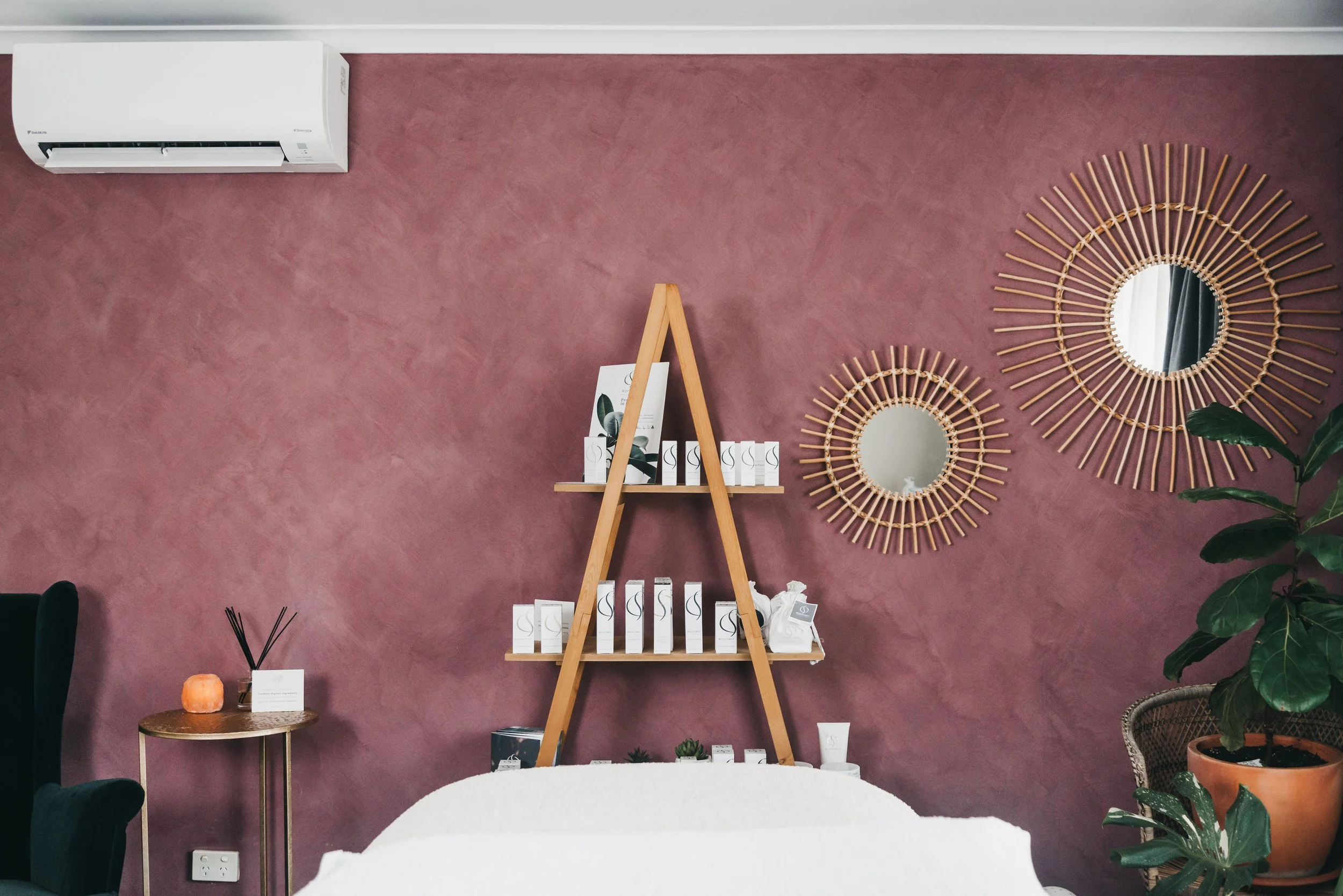 Decorative interior wall with two round starburst mirrors, a wooden ladder shelf with products, a potted plant, and an air conditioner.