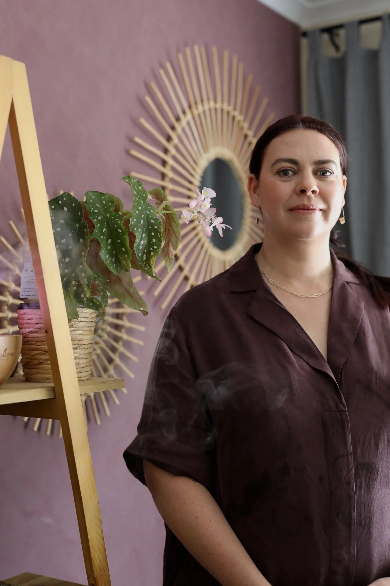 A woman with dark hair and light skin standing indoors in front of a pink wall with a sunburst mirror. She is wearing a dark brown shirt and a gold chain necklace, with a small earring visible. To her left, there is a wooden shelf with a potted plant with large green leaves and pink flowers.