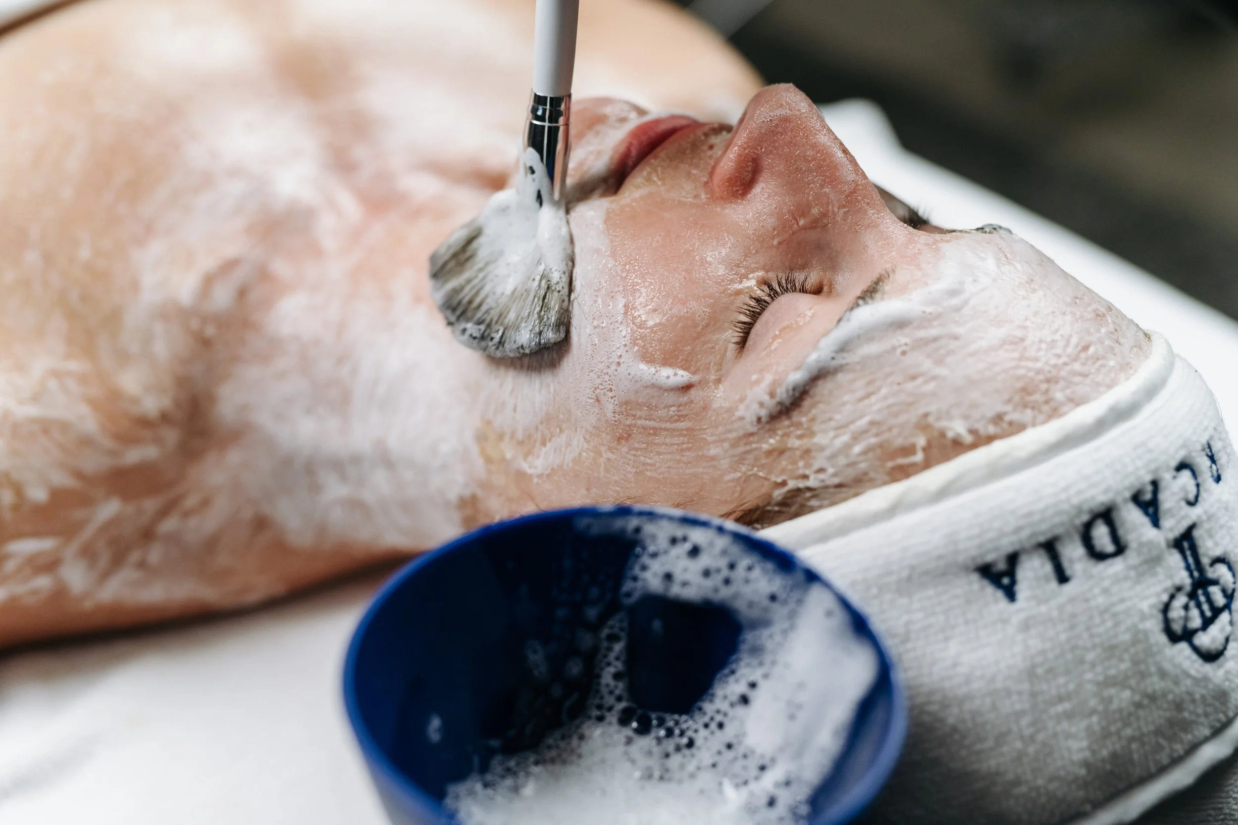 A woman receives a facial treatment, with foamy soap on her face and her eyes closed, lying on a treatment bed.