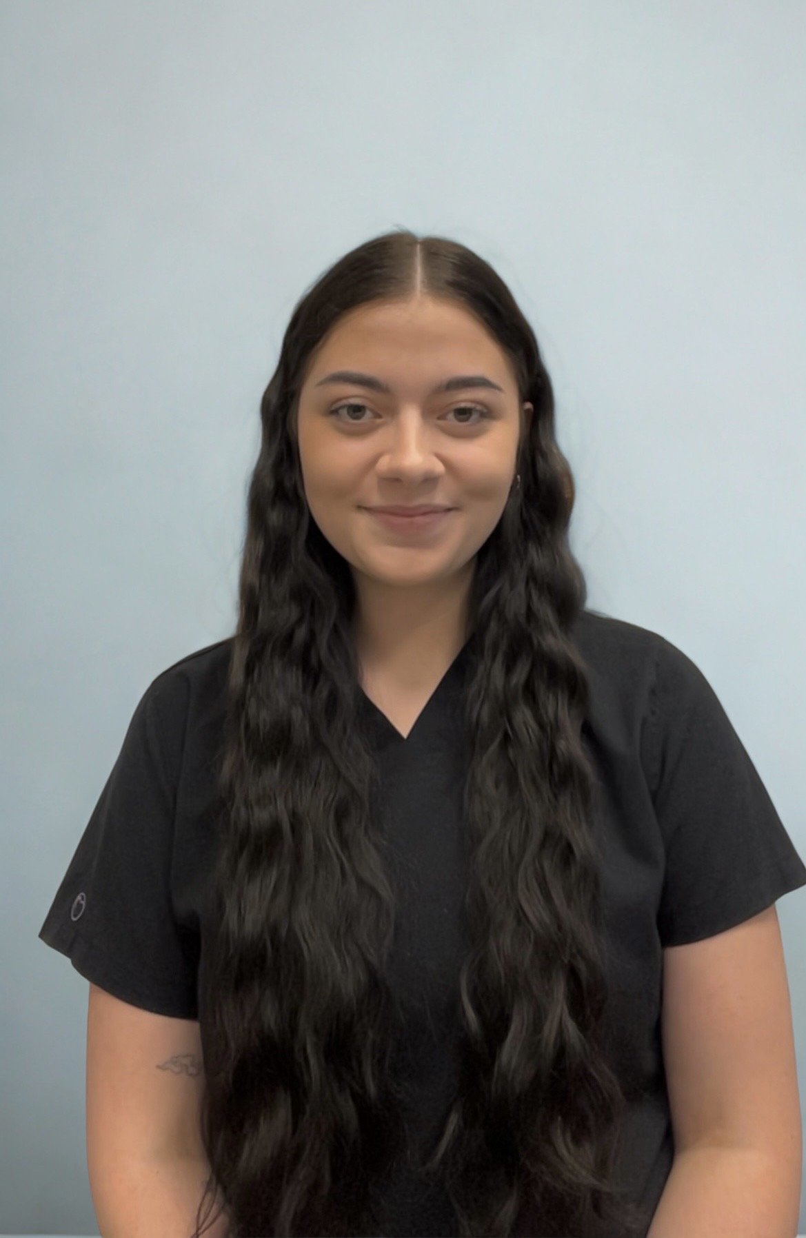 A woman with long, wavy dark hair and light skin, wearing a black shirt, smiling softly, standing against a plain light blue wall.