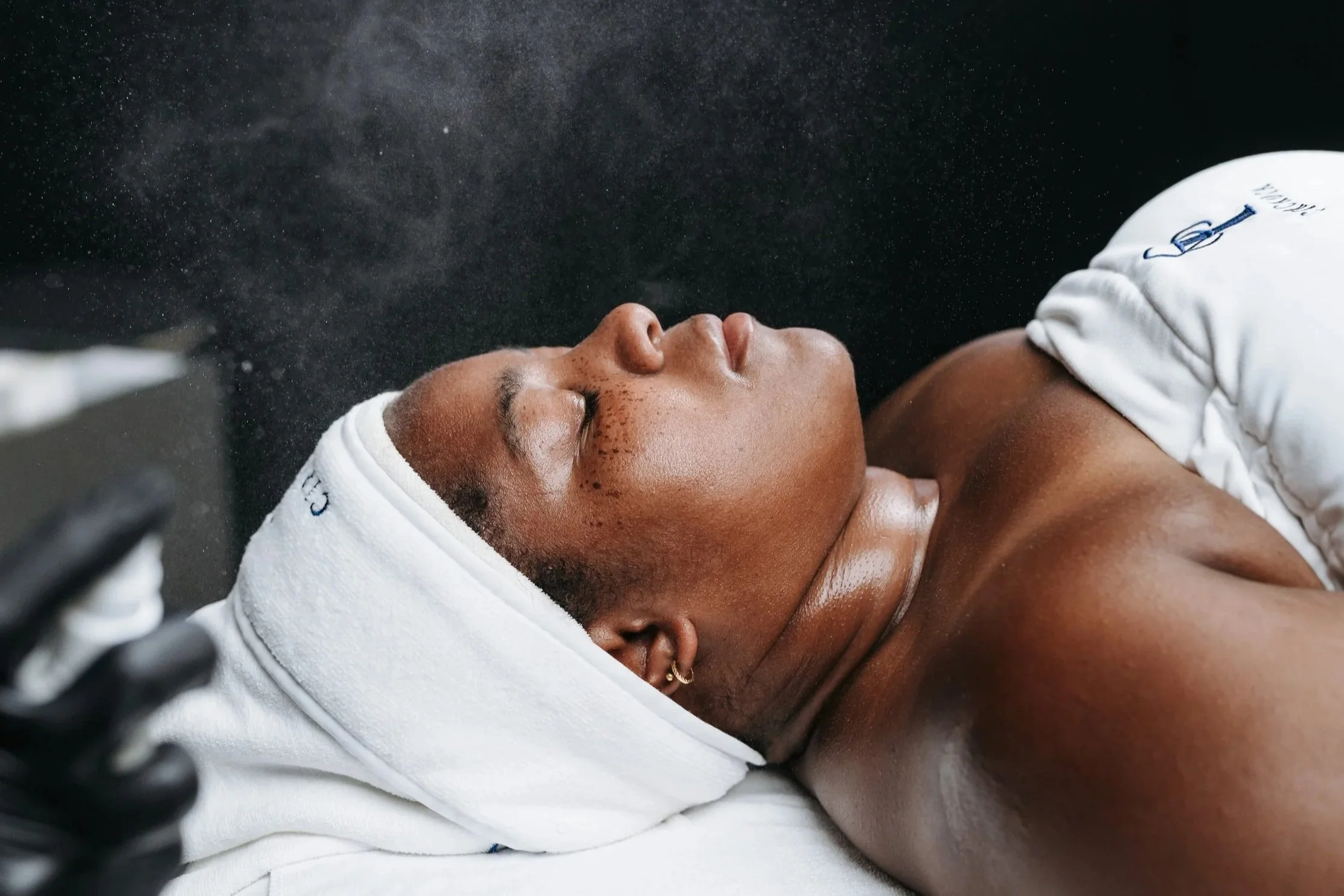 A person lying on a treatment bed with a white towel on their head, receiving a facial treatment amidst a fine mist, in a spa or skincare clinic.