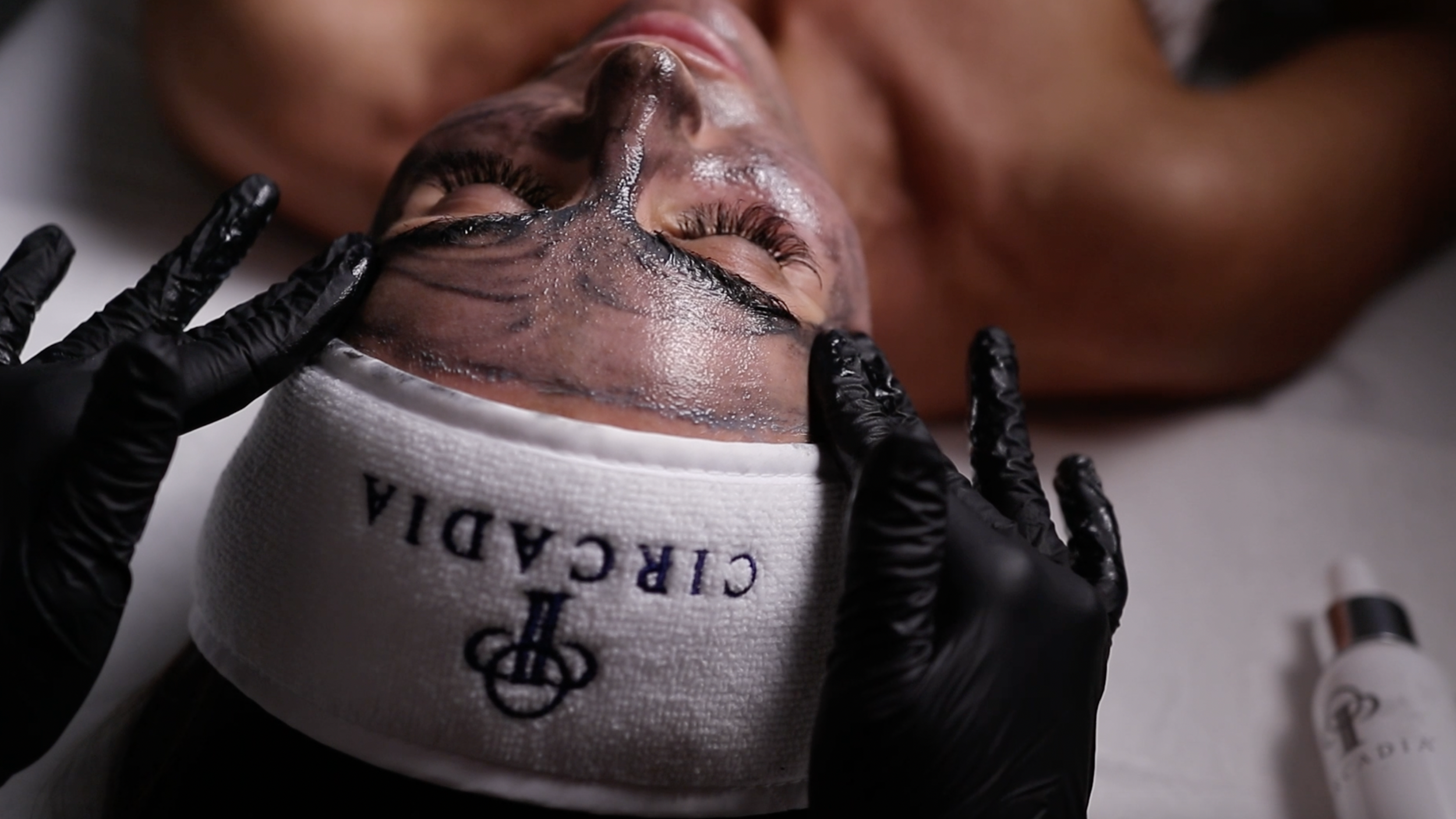 A person lying down with eyes closed wearing a white headband and receiving a facial treatment with black gloves applying a product to their face.