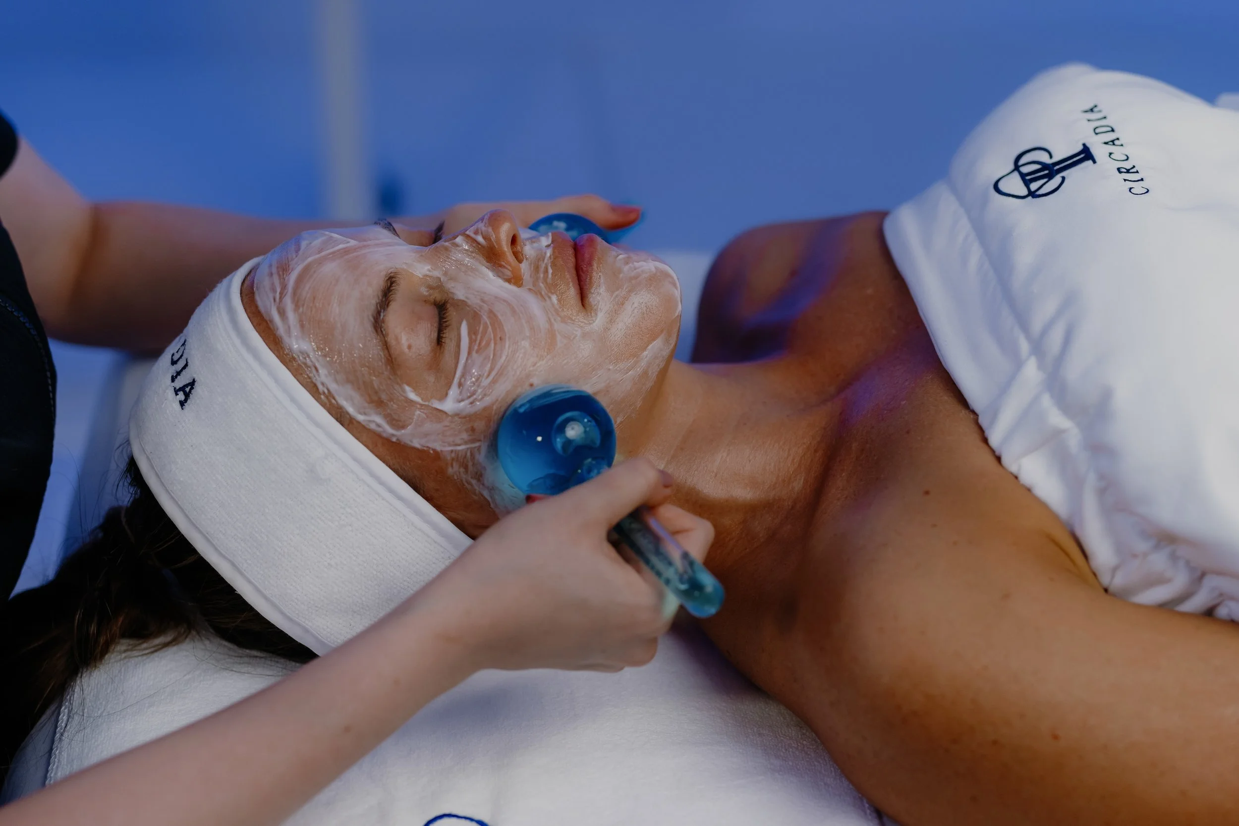 A woman with her eyes closed, lying on a treatment table with a white headband that reads 'ELEMIS', receiving a facial treatment. A professional is gently applying a facial mask or cream to her face, and she is wearing blue ear covers. The background is a solid blue color.