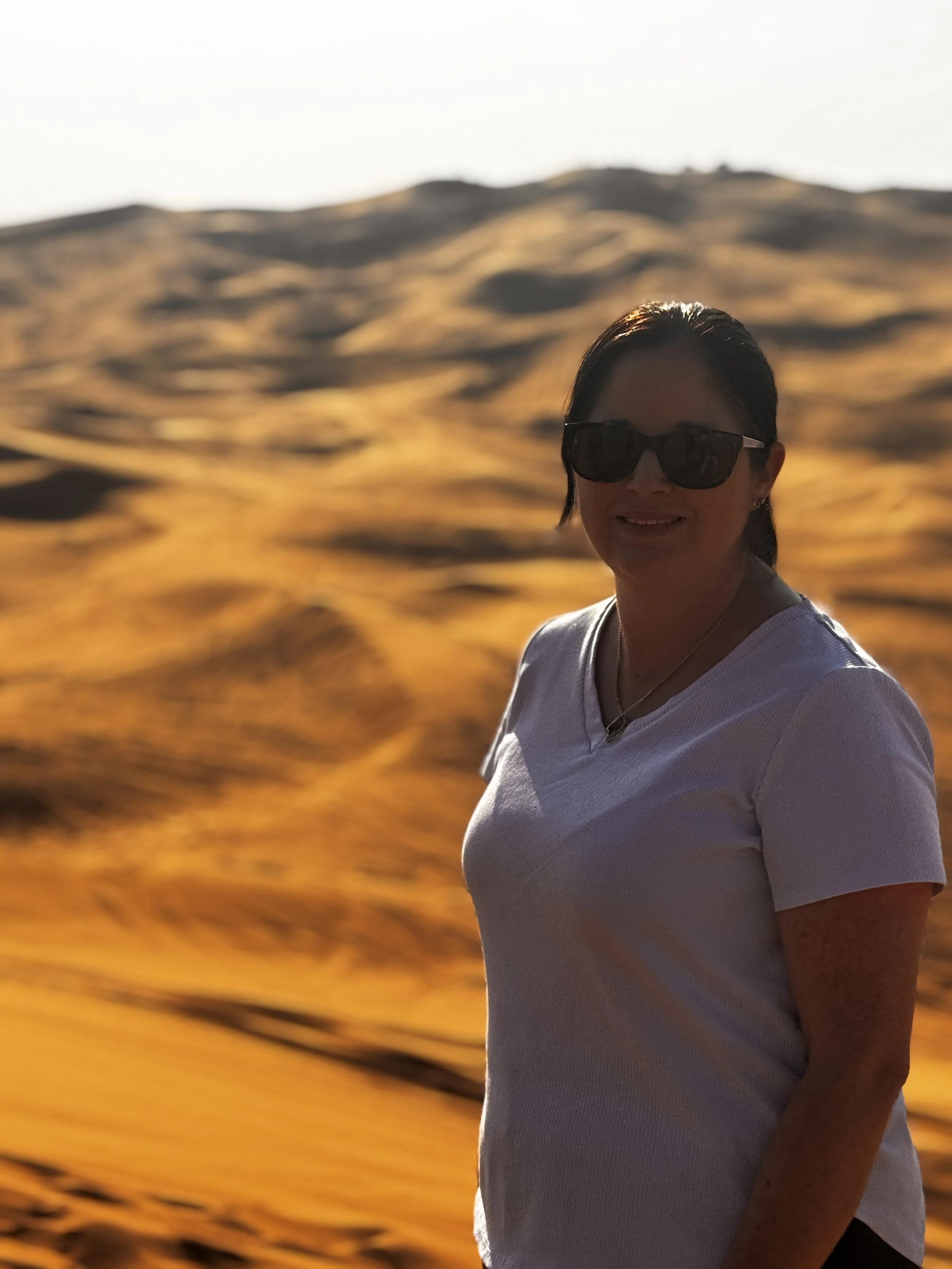 Woman wearing sunglasses and a white t-shirt standing outdoors during sunset with a landscape of rolling hills in the background.