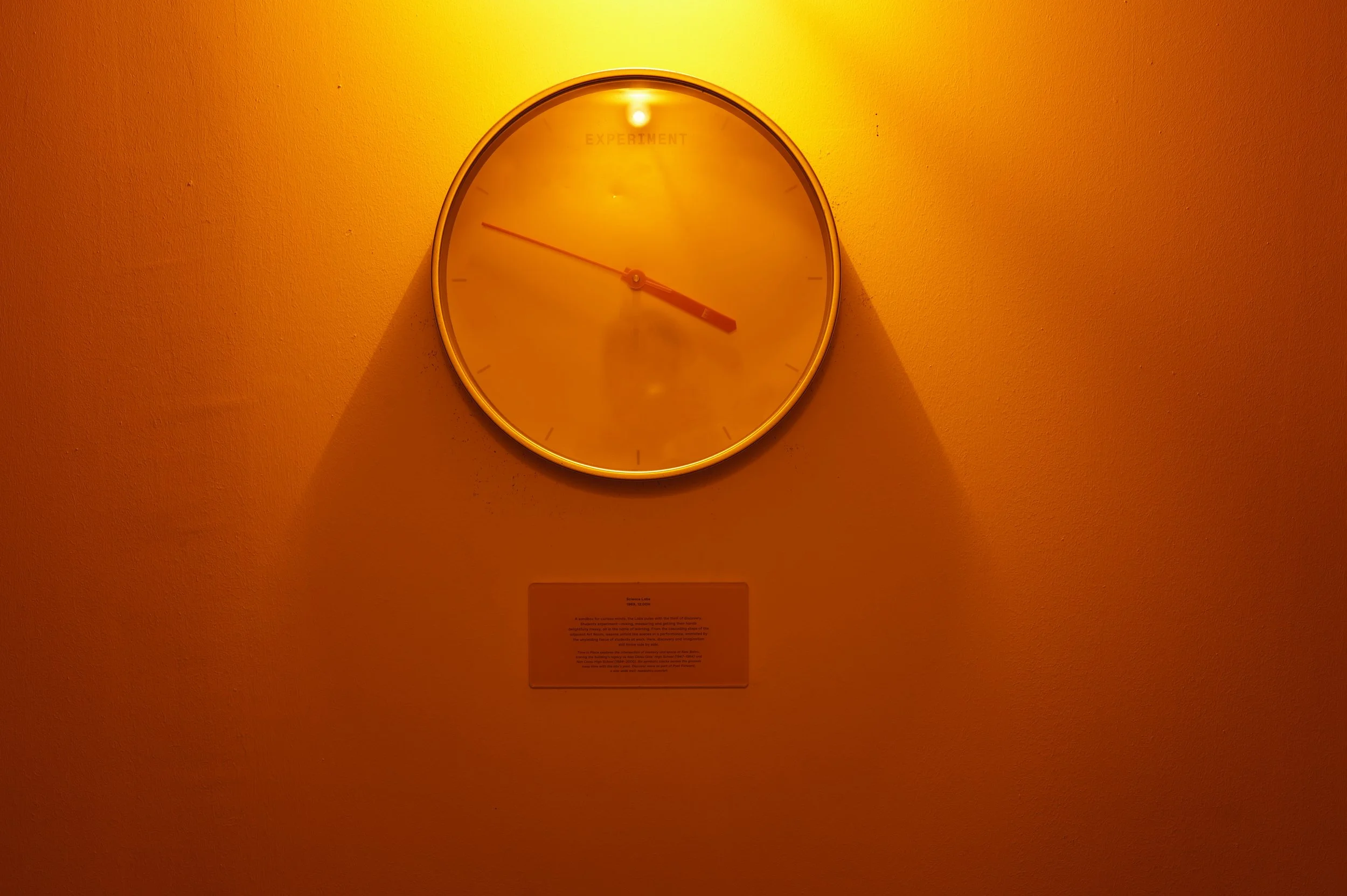 A round wall clock with a white frame, showing the time as approximately 3:20, is mounted on a wall illuminated with a yellow-orange light. There is a small label or plaque below the clock.