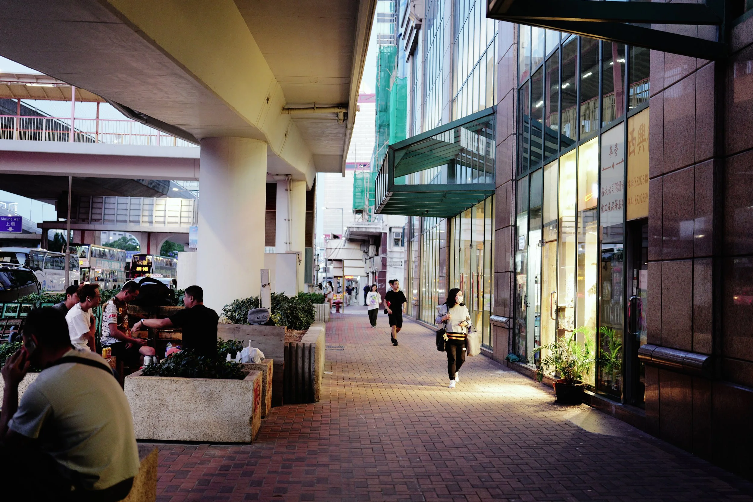 Urban street scene with pedestrians walking and sitting; modern glass-front buildings; highway overpass overhead; vehicles parked to the side; sunlight illuminating the area.