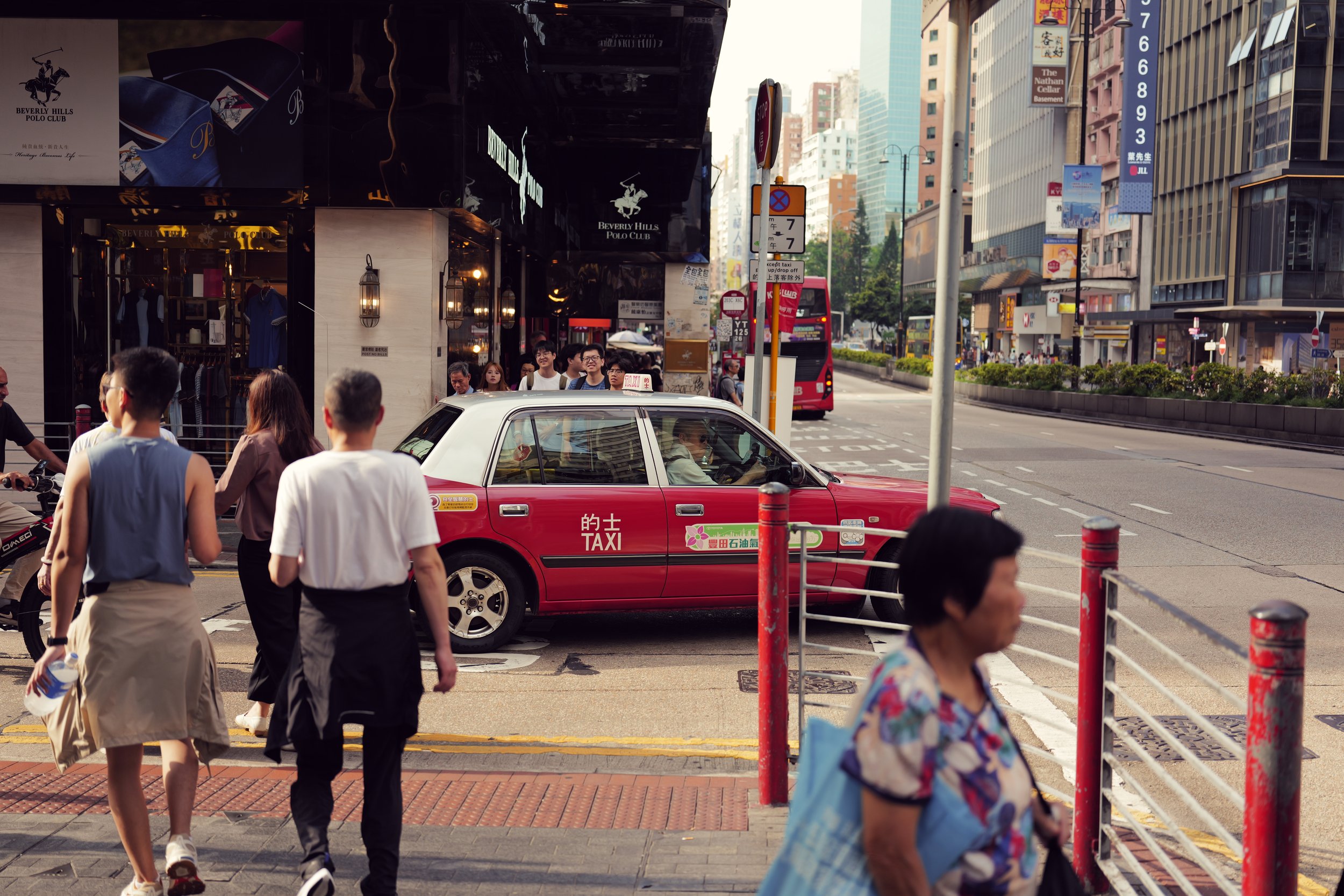 People crossing a busy city street with tall buildings, taxis, and buses visible. A red taxi with Chinese characters on the side is in the foreground.
