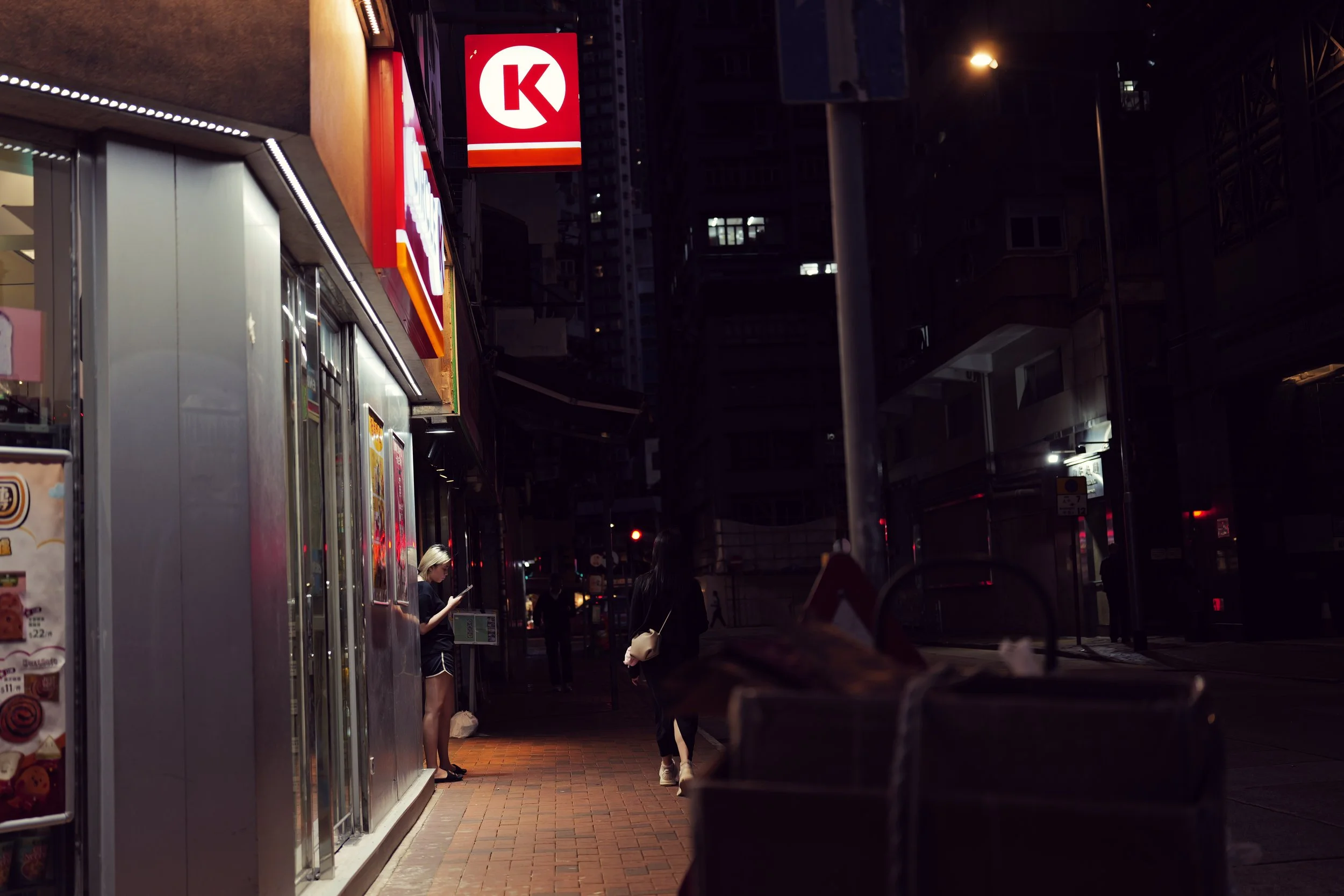 Nighttime scene of a city street with people standing outside a store, illuminated signs, and tall buildings in the background.