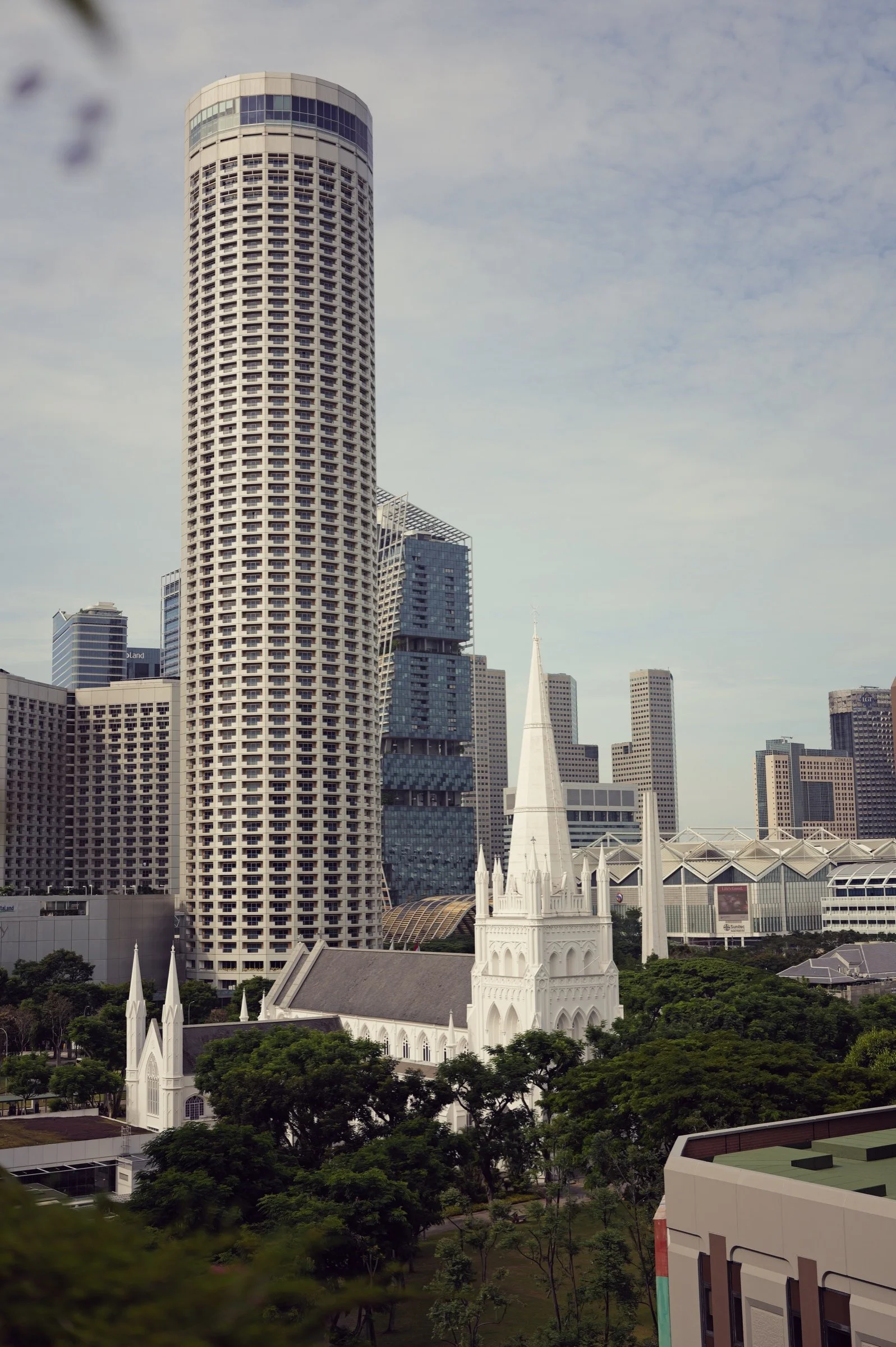 A tall rounded tower rises above trees and neighbouring city buildings beneath a pale Singapore sky.