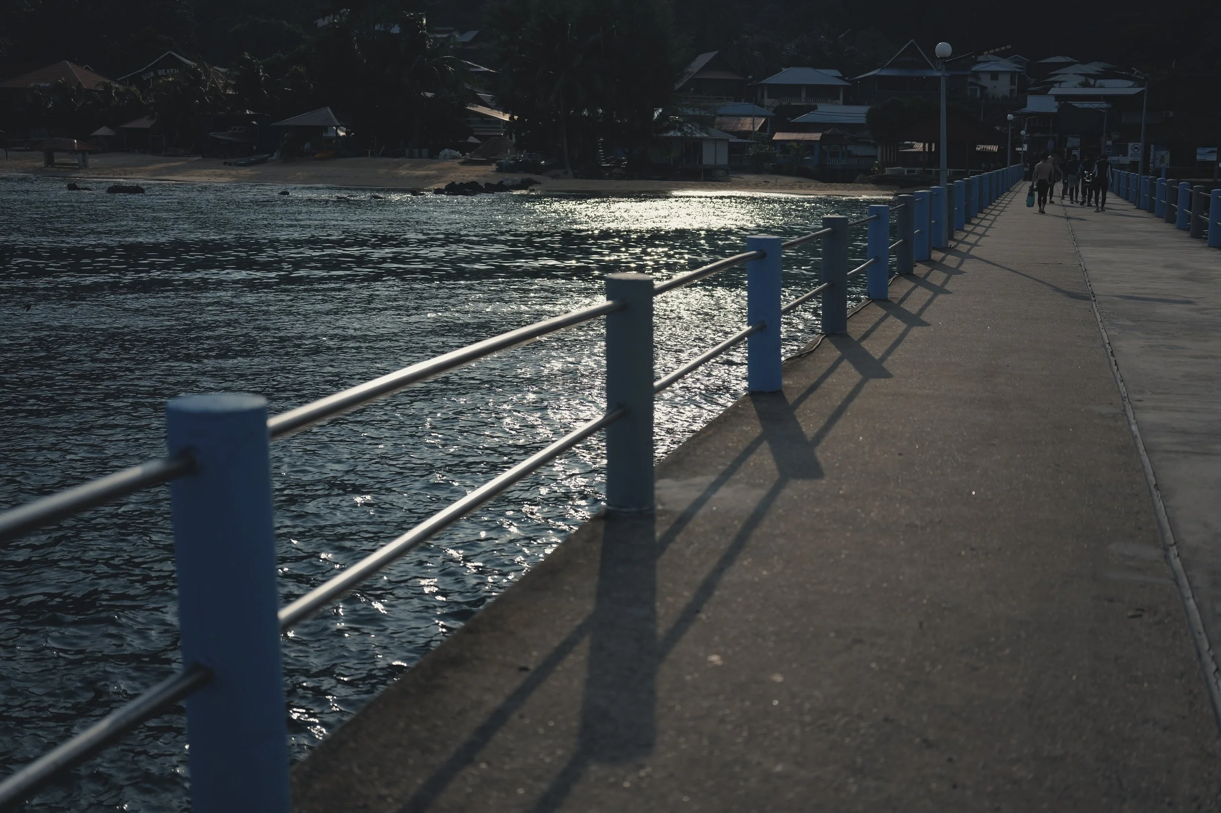 A concrete pier with metal railings extends over the water near a small town or village at dusk or dawn. Houses and palm trees are visible along the shoreline in the background, and a few people are walking along the pier, some with shopping bags or 