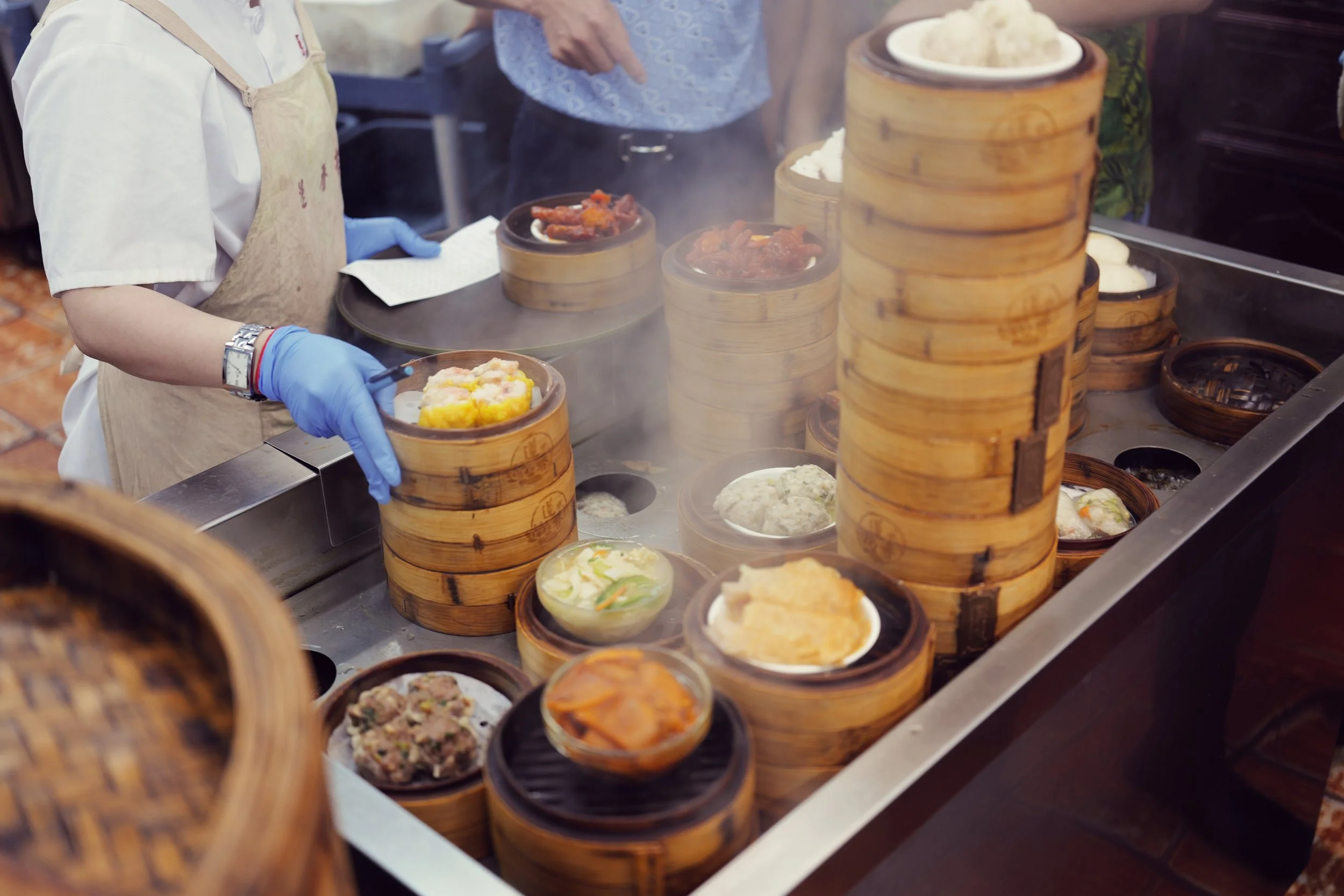 A person wearing a beige apron and blue gloves is serving dim sum in small bamboo steamers and bowls at a food stall. The steamers contain various types of dim sum, and other dishes are visible on the table.