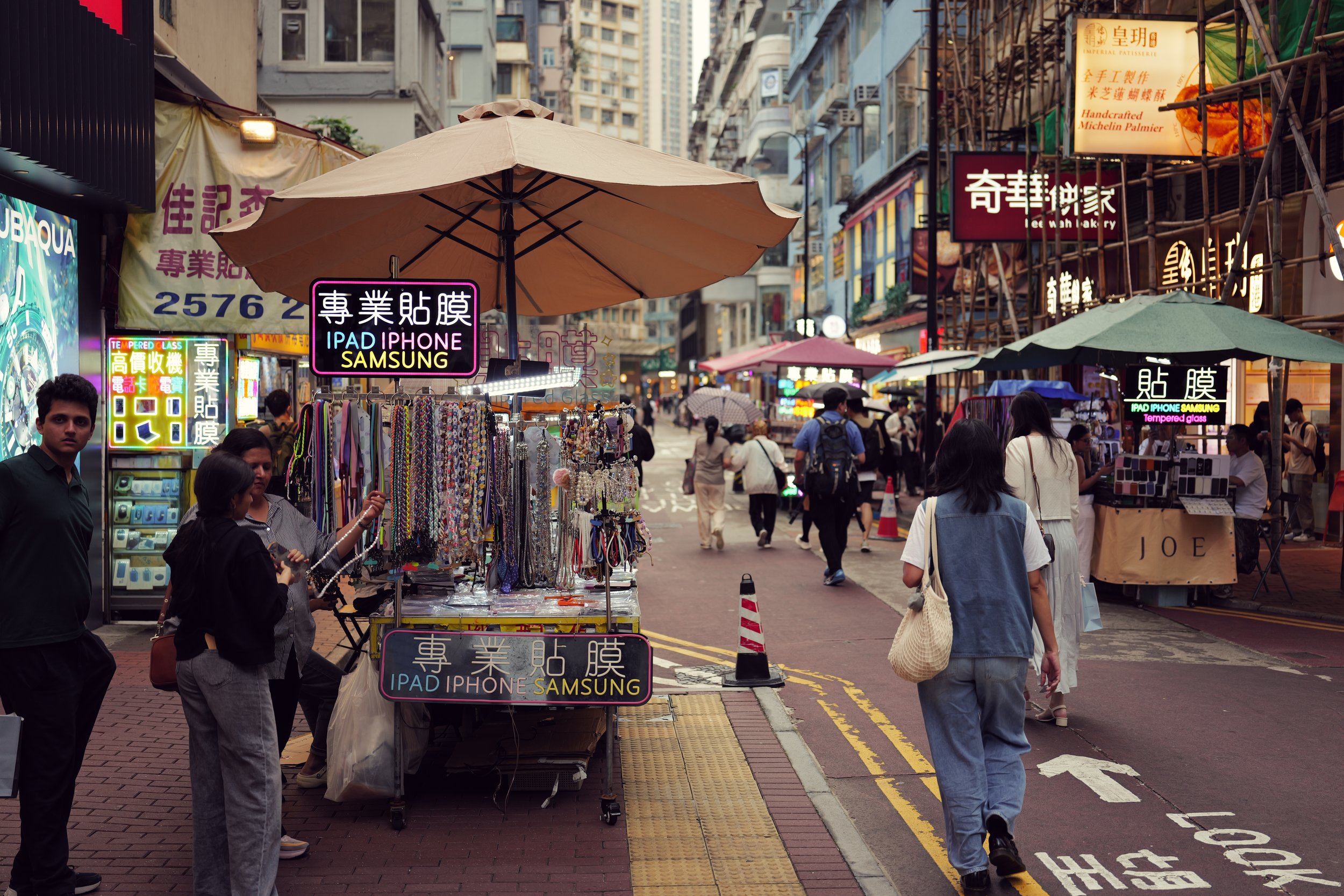 A busy street market with shop stalls, umbrellas, and people walking. Signs in Chinese advertising electronic accessories and baked goods are visible, along with a yellow traffic line and a pedestrian crosswalk.