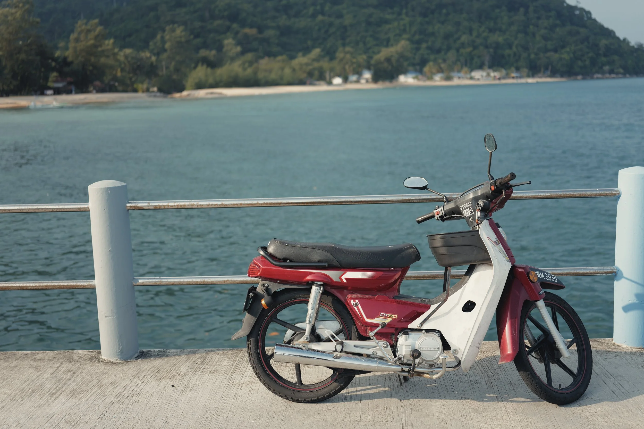 Red and white electric scooter parked by a water body, with hills and houses in the background, and a metal railing in the foreground.
