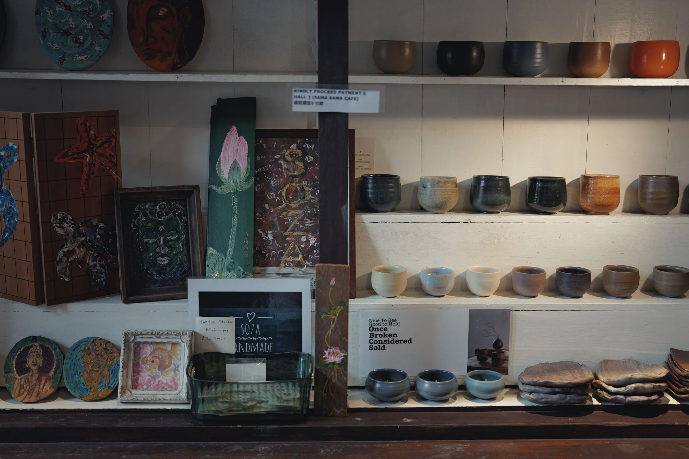 Ceramic cups and framed paintings fill a sunlit wooden shelf.