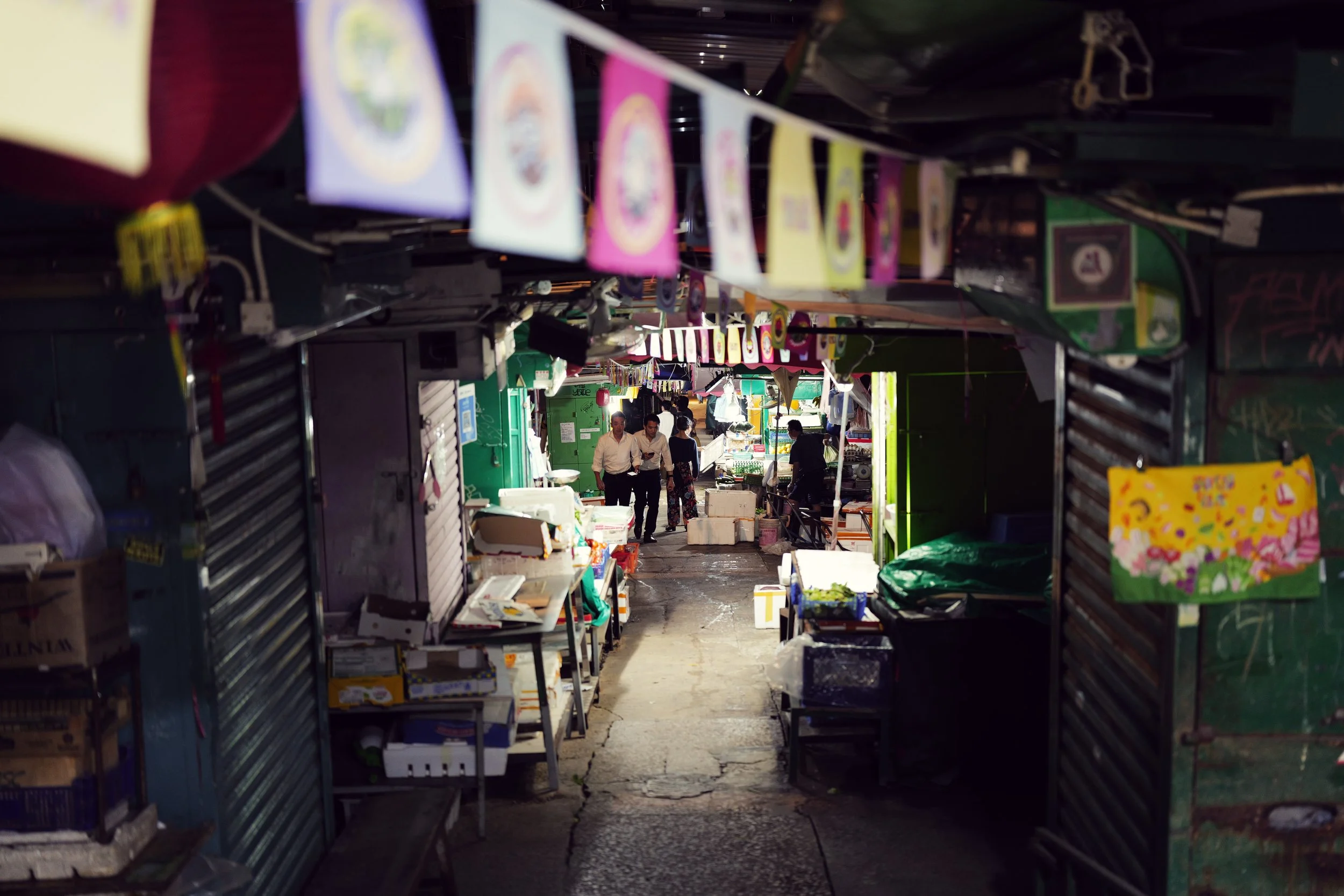 An outdoor marketplace with stalls on both sides and people walking through, viewed from a narrow alley with overhead colorful banners.