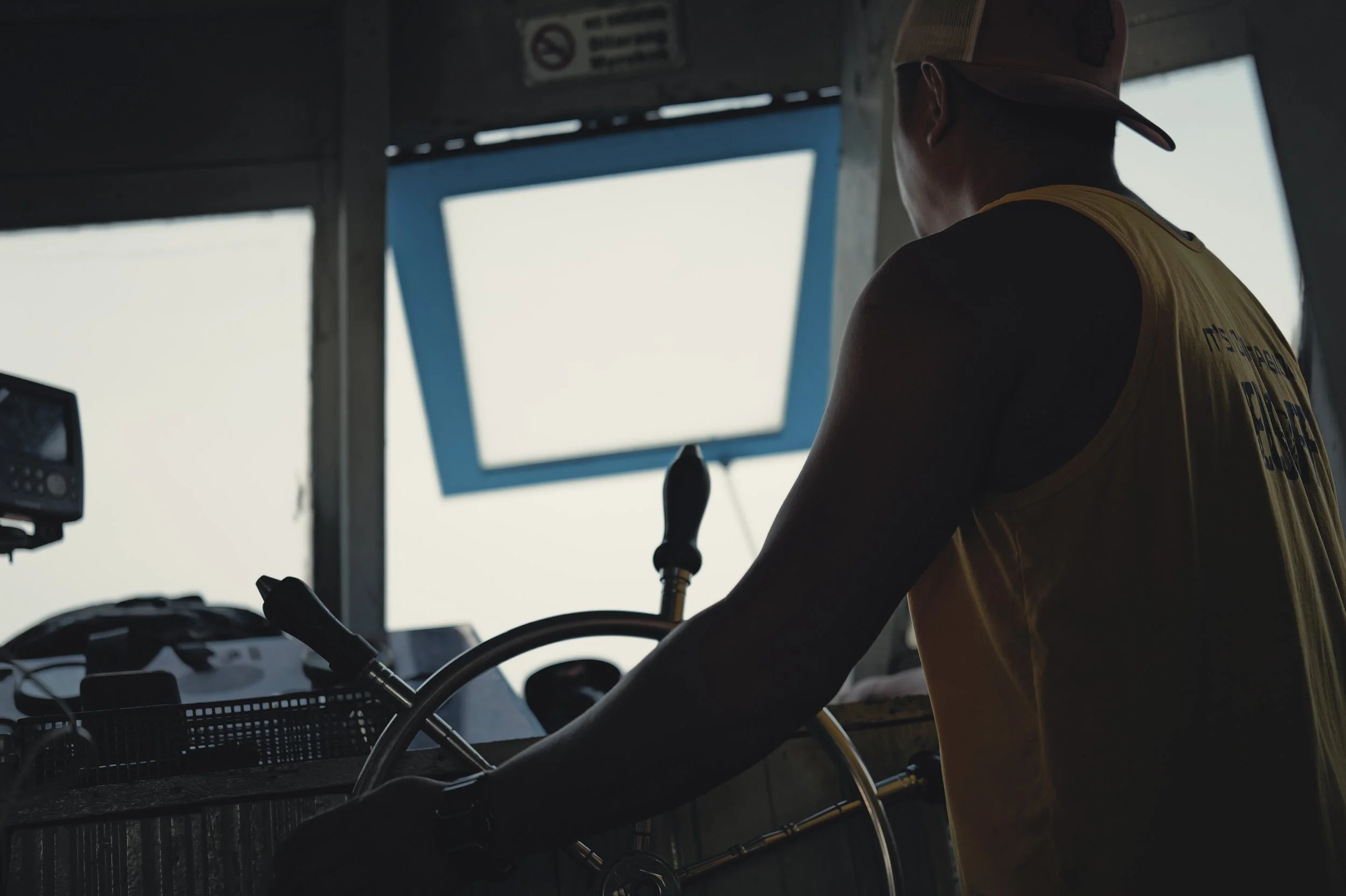 A person in a yellow safety vest and camouflage cap operating a vehicle or machinery inside a dimly lit structure, with windows showing a bright sky outside.