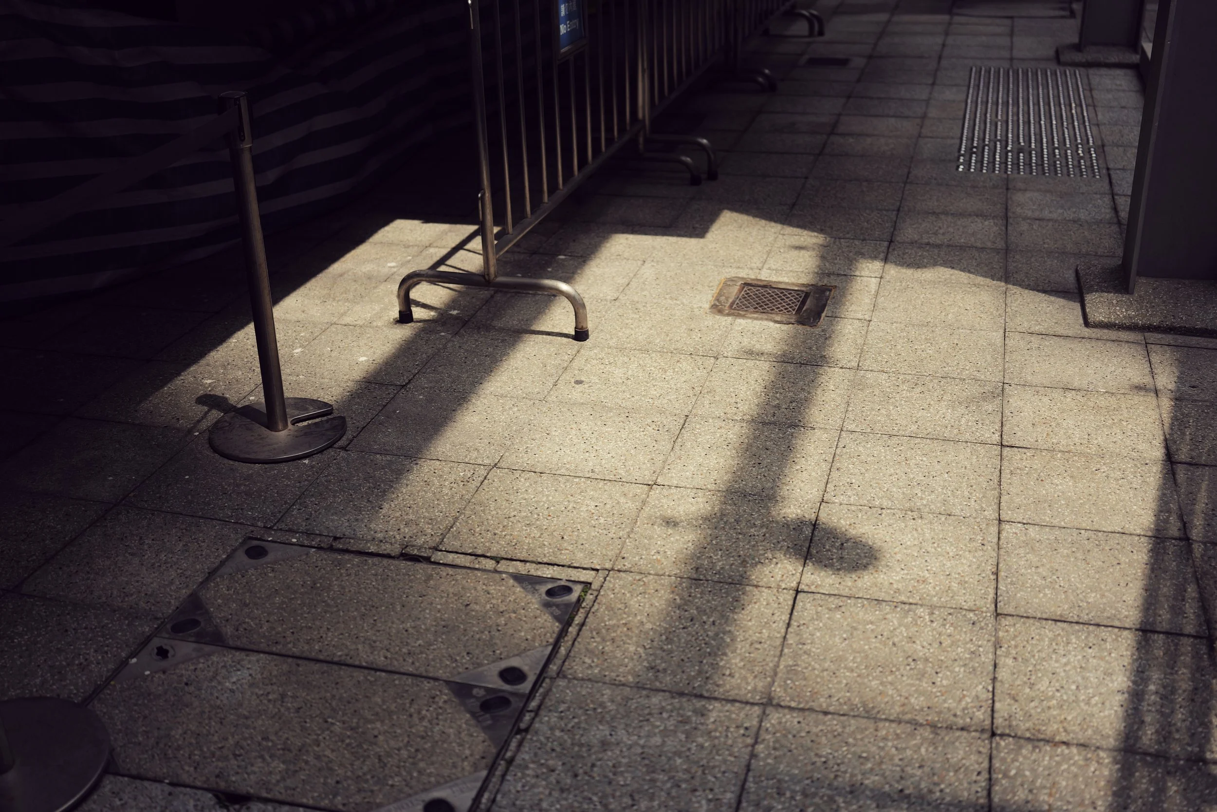Empty sidewalk area with metal barricades and a drain grate, casting shadows from nearby structures.