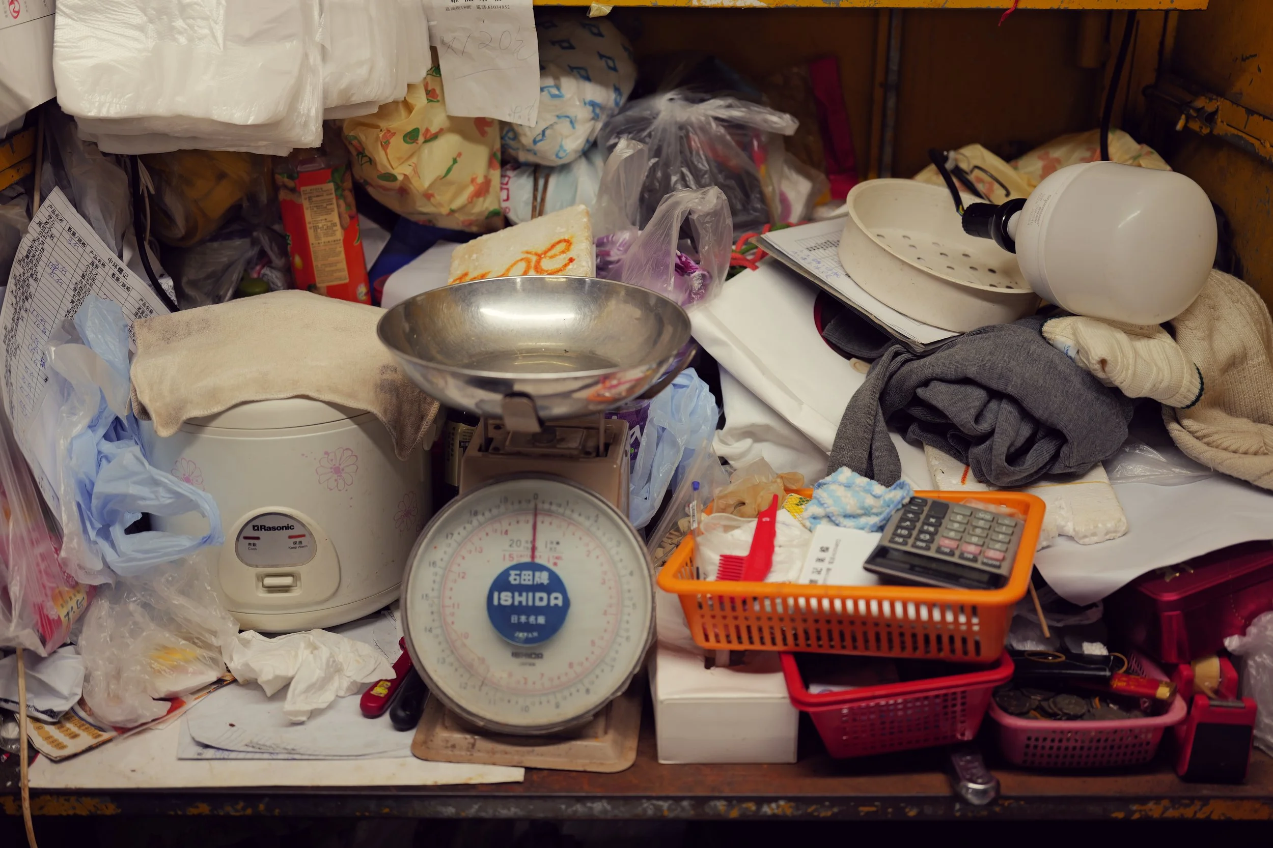 Cluttered work desk with various objects including a white humidifier, plastic bags, a vintage scale, a calculator, a trash bin, and miscellaneous papers and items.