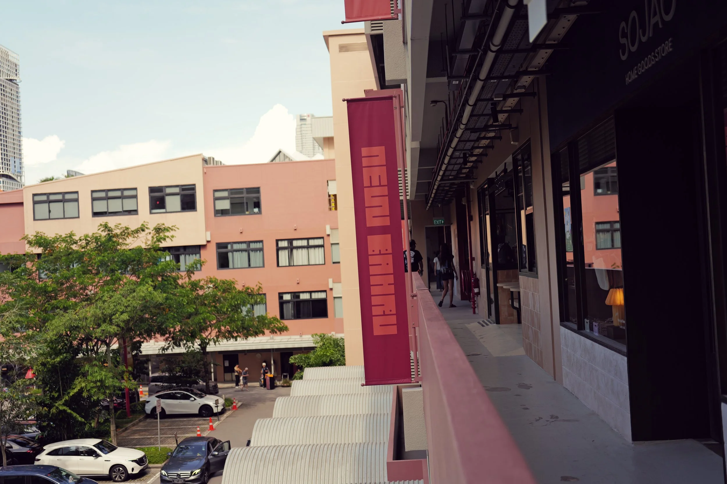 A view of a pink building with several windows, trees, a parking lot with cars, and a sign that reads 'LITTLE ITI' on a balcony railing. There are people walking on the balcony of the building.