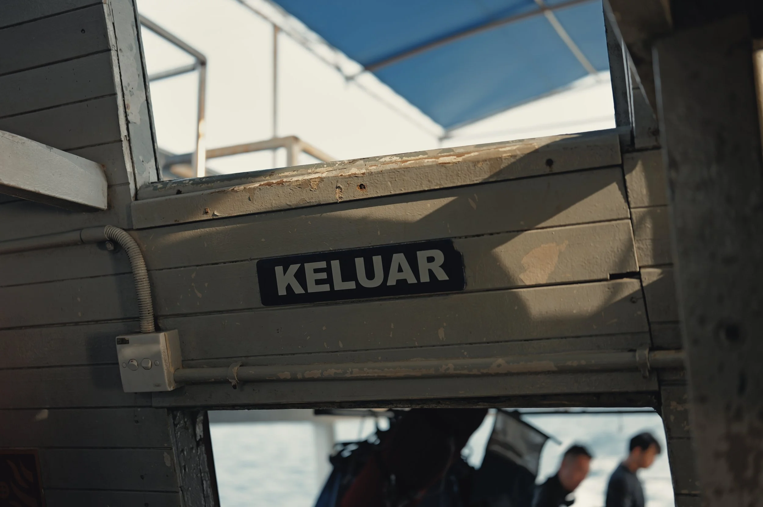 Close-up of a building with a sign that reads 'KELUAR', an electrical switch, and several people in the background beneath a blue roof.