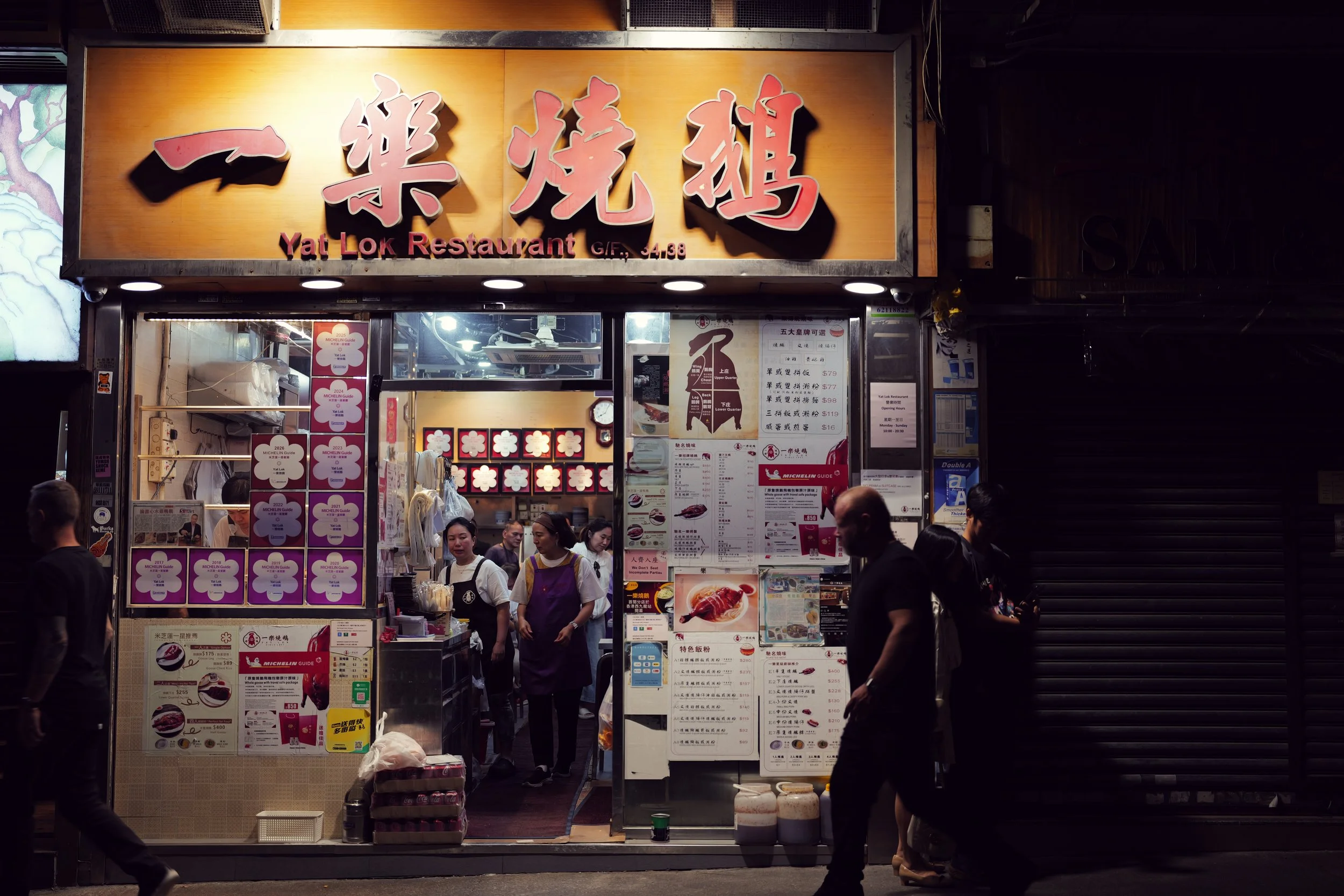 Night scene of a Chinese restaurant storefront with large pink Chinese characters and smaller English text reading 'Yat Lok Restaurant'. The storefront has menu boards, posters, and people dining inside and outside.